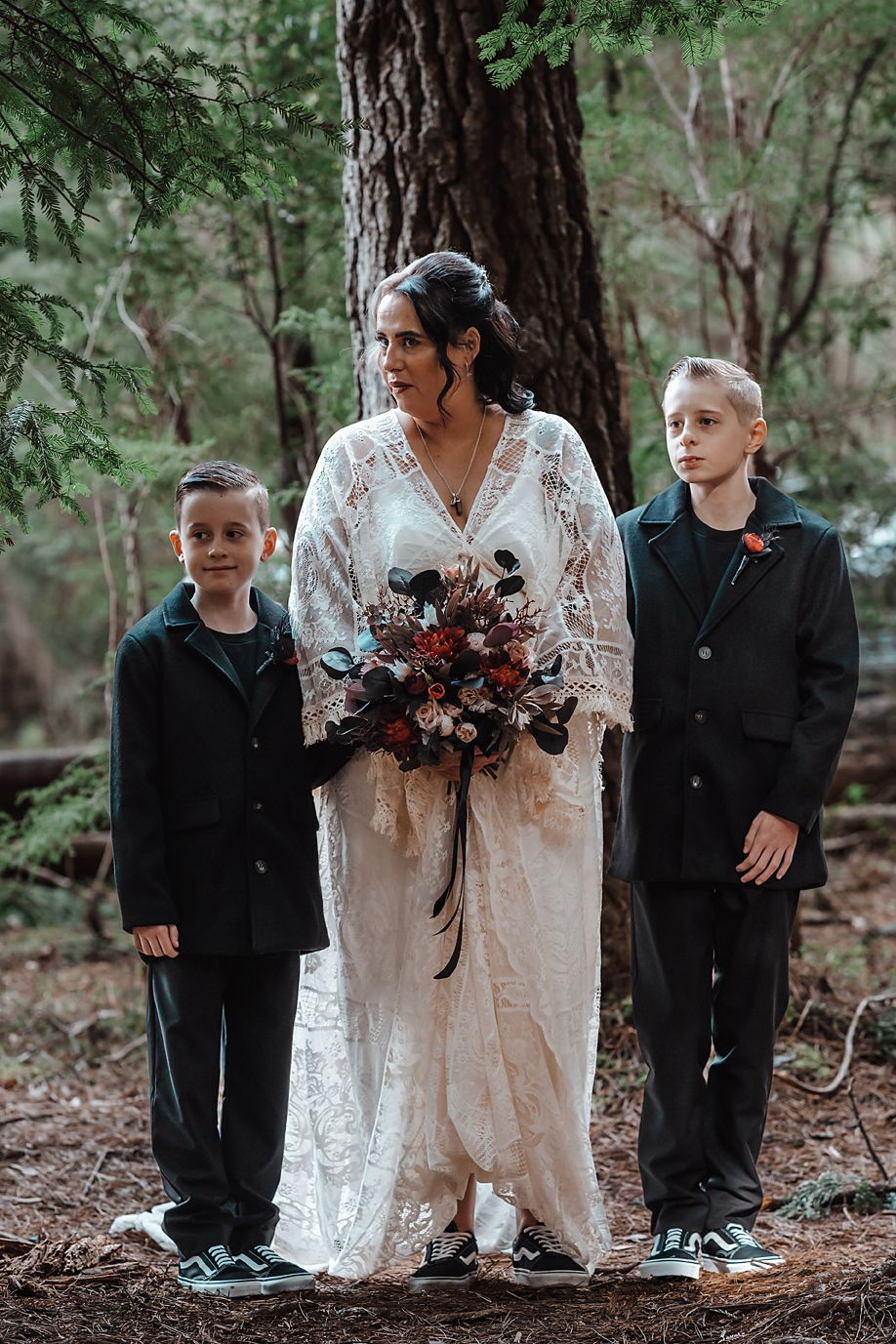 Intimate forest wedding scene showing bride in lace gown surrounded by attendants in black attire.