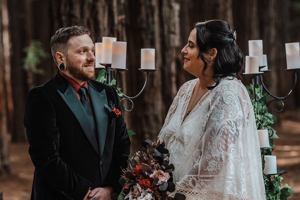 Two people in formal wedding attire share a moment in a forest with elegant candelabras in the background.