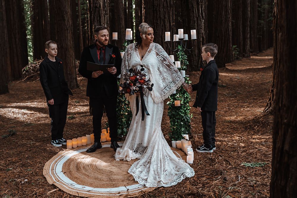 A wedding party gathers for photos among tall trees in a rustic forest setting.
