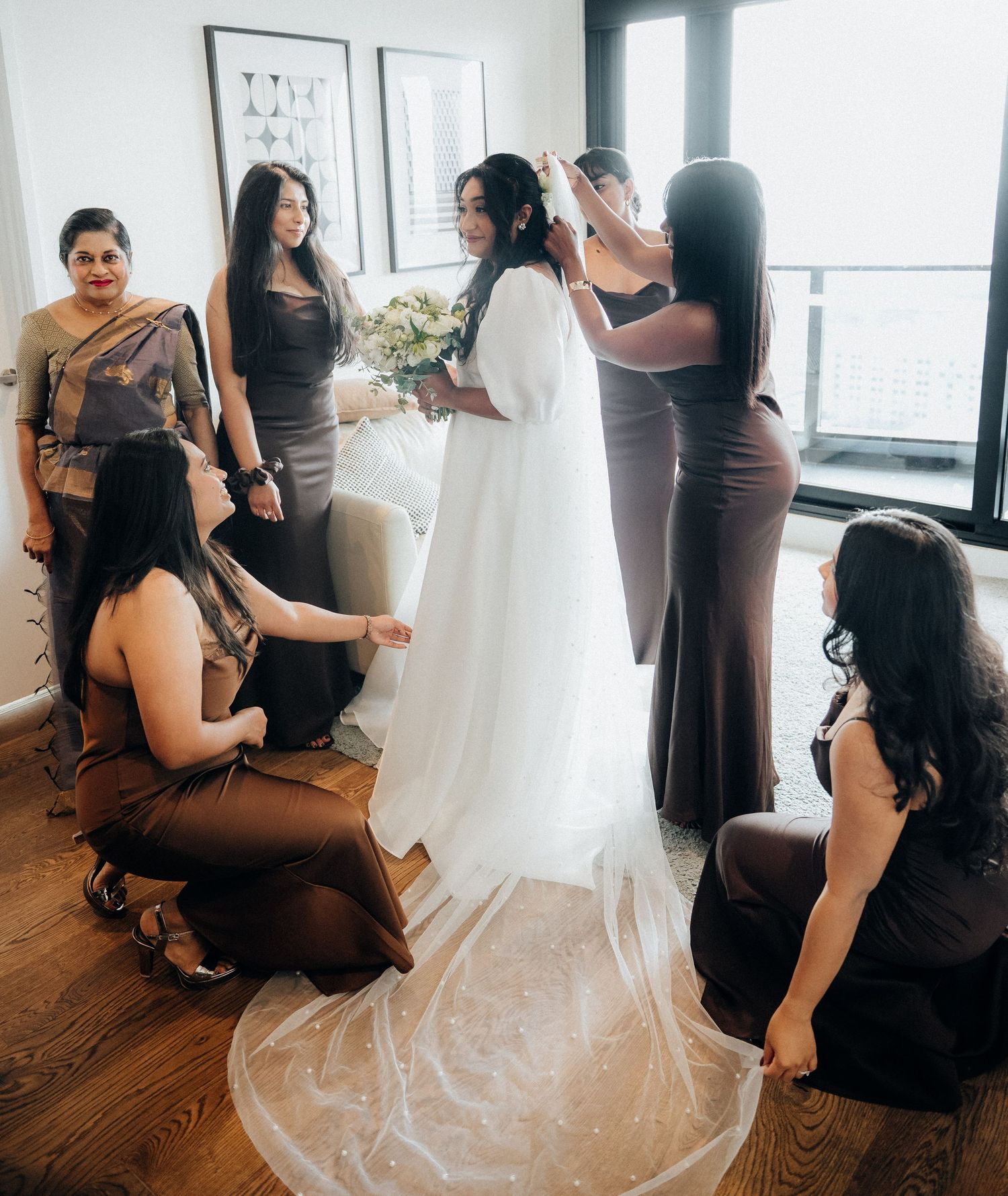 Several bridesmaids in dark dresses help the bride prepare her wedding gown in a bright room with large windows.