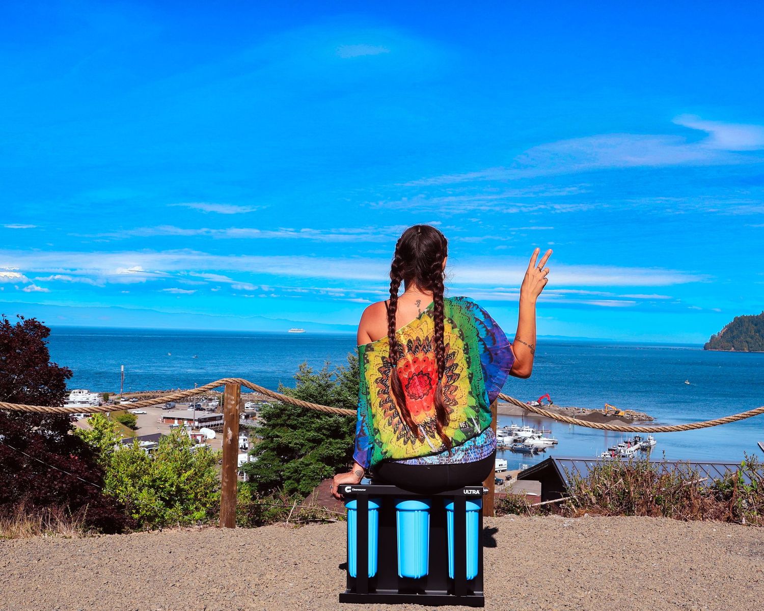 Woman in tie-dye shirt sitting on cooler enjoying ocean view on sunny day.