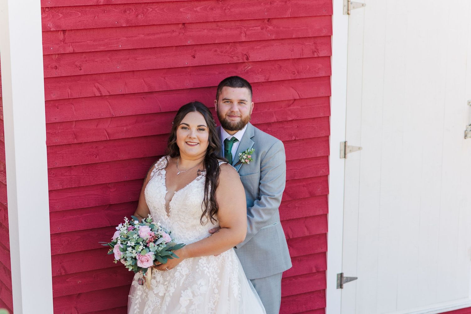 A couple poses together on their wedding day against a bright red barn wall while holding a beautiful floral bouquet.