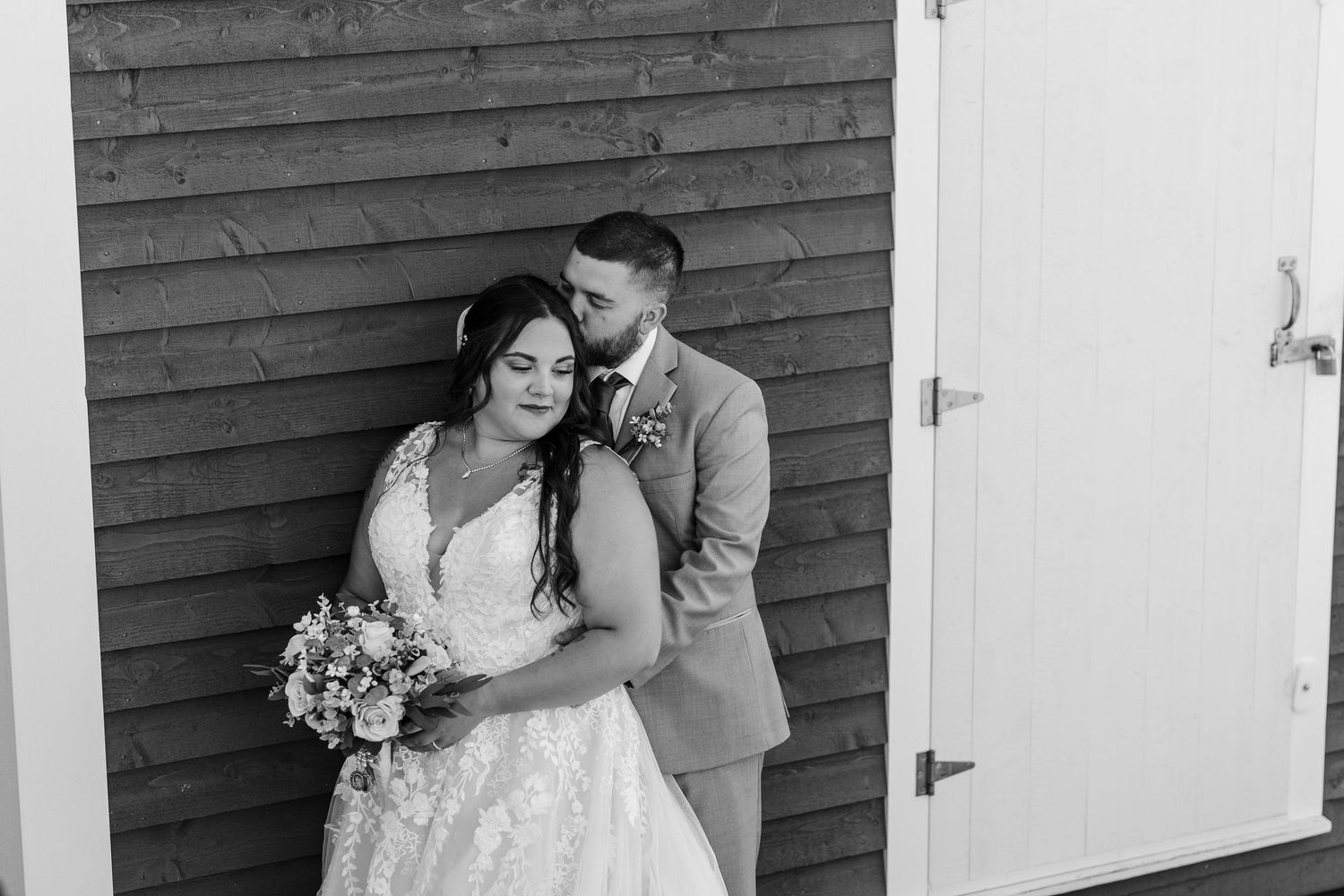 Black and white photo of a romantic wedding couple embracing against a rustic wooden wall with bride holding a bouquet.