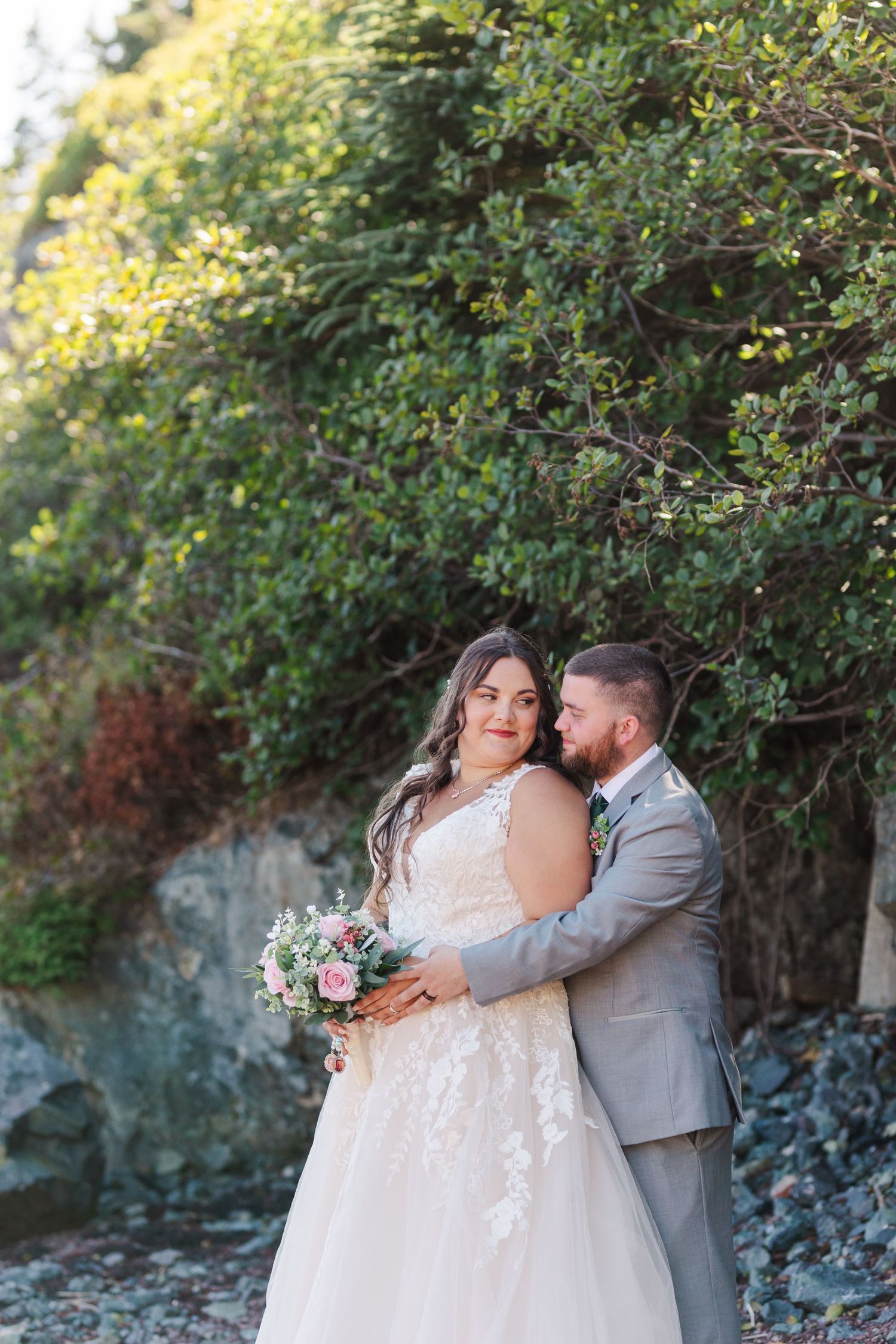 A couple embraces during their wedding photoshoot in a lush outdoor setting with trees and rocks in the background.