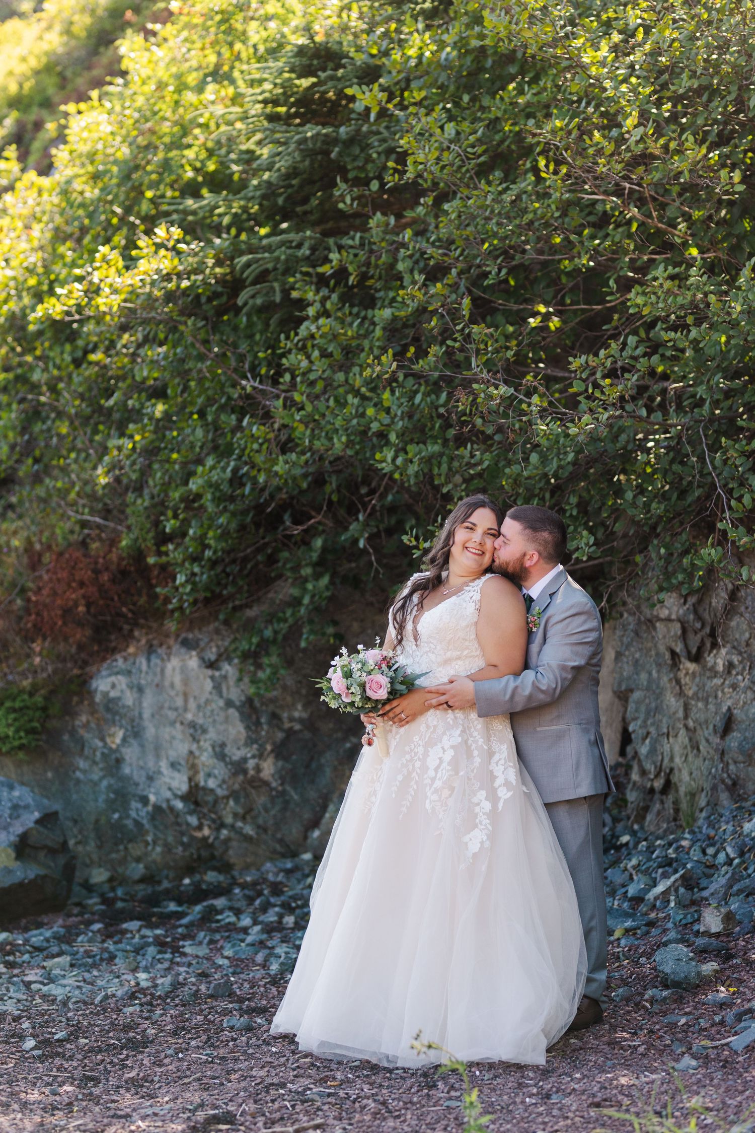 Newlywed couple shares romantic kisses in front of greenery and stone wall in wedding attire.