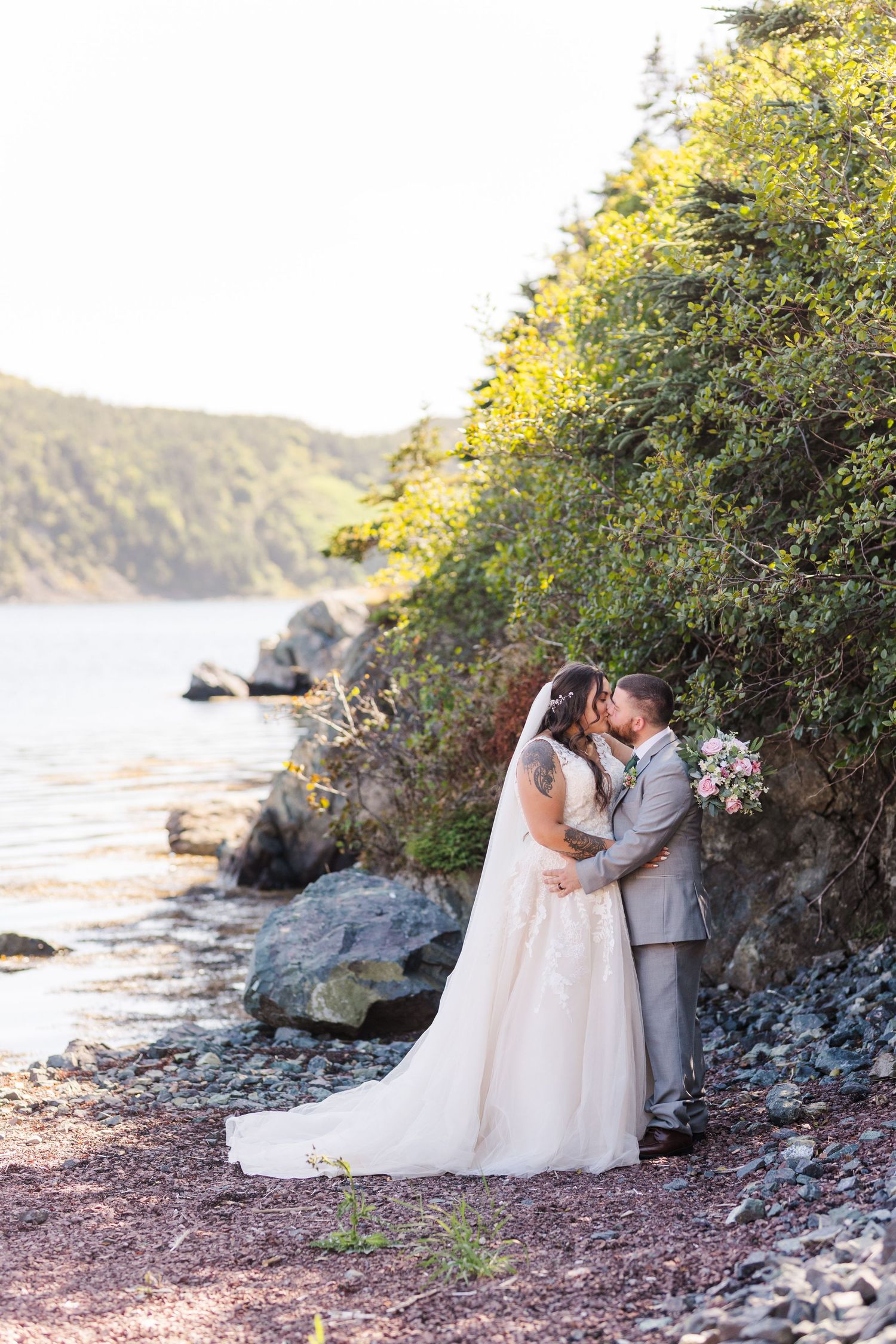 A couple shares romantic moments near a rocky coastline at sunset with vibrant foliage in the background.