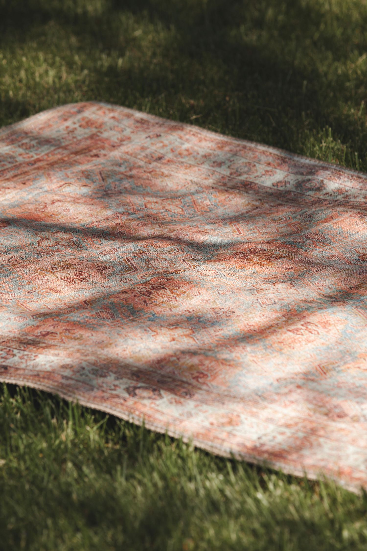 A patterned blanket with reddish tones lies on grass with dappled sunlight and shadows falling across its surface.
