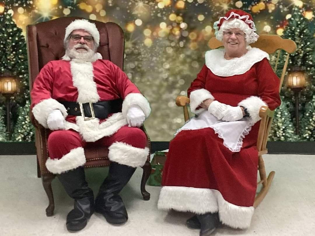 Santa Claus figures sit on chairs near decorated Christmas trees with twinkling lights creating a festive holiday scene.
