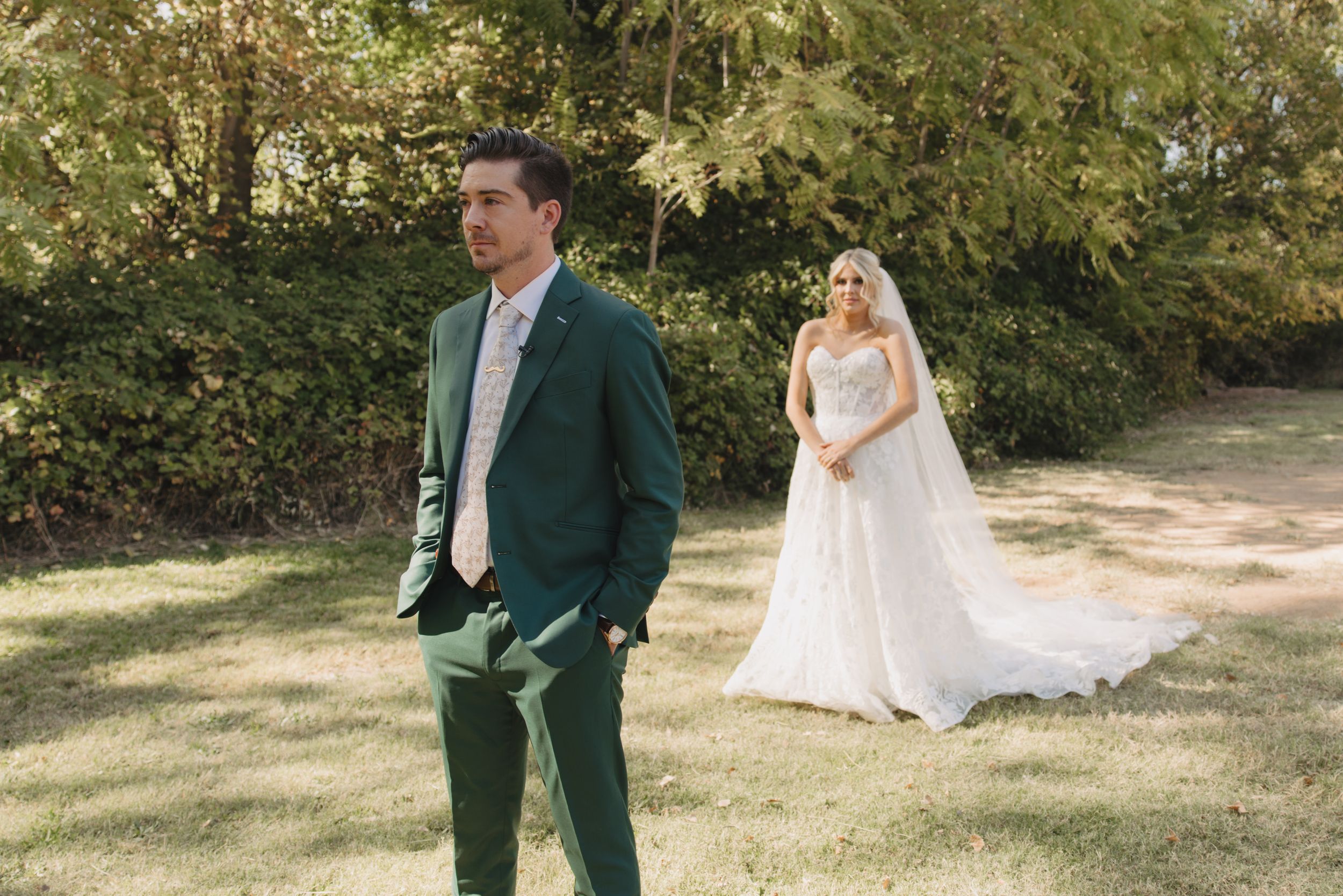 A couple in wedding attire pose in a sun-dappled garden with the bride wearing a flowing white gown.