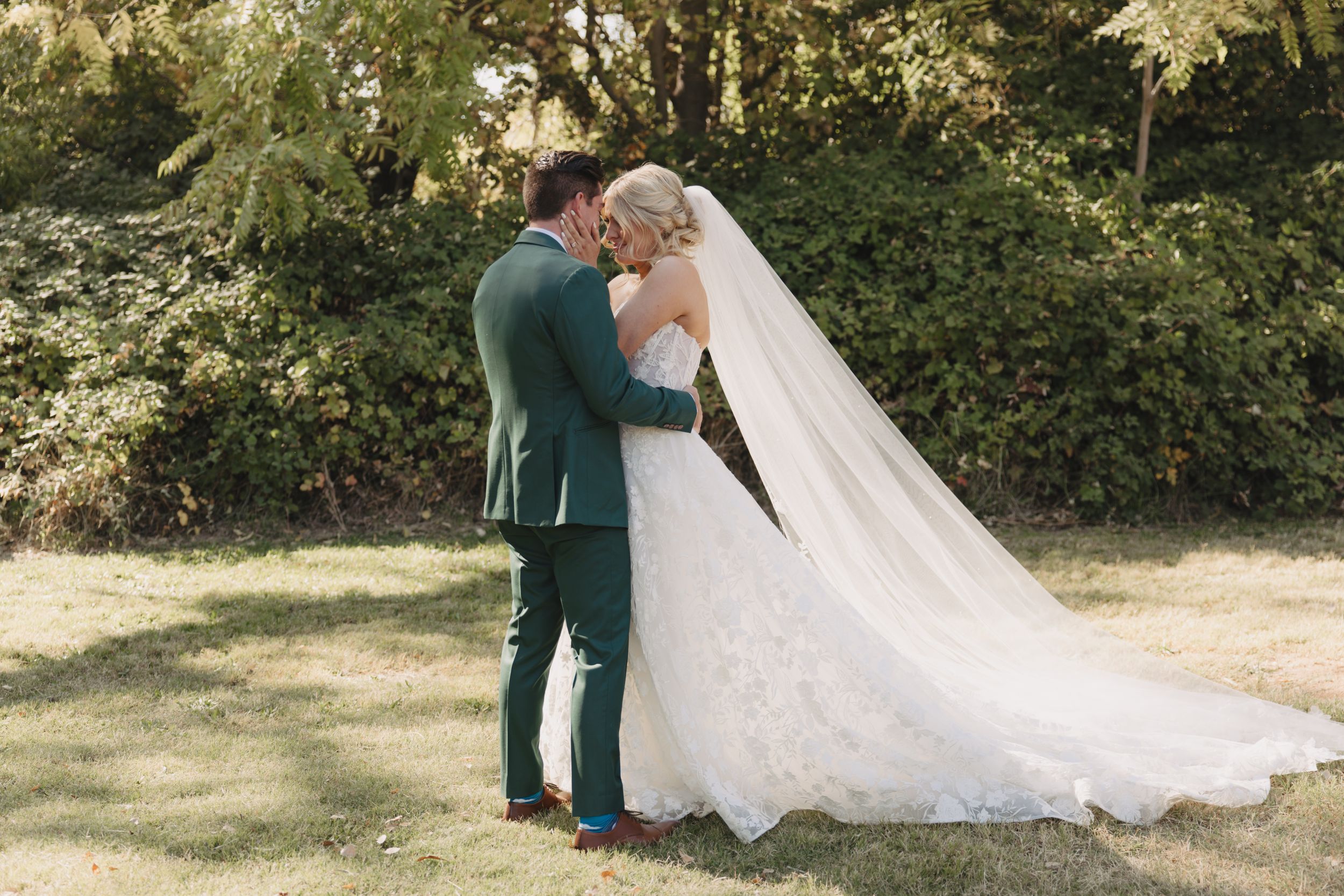 A couple in wedding attire shares a romantic moment on a sunny lawn with a long white flowing dress trailing behind them.