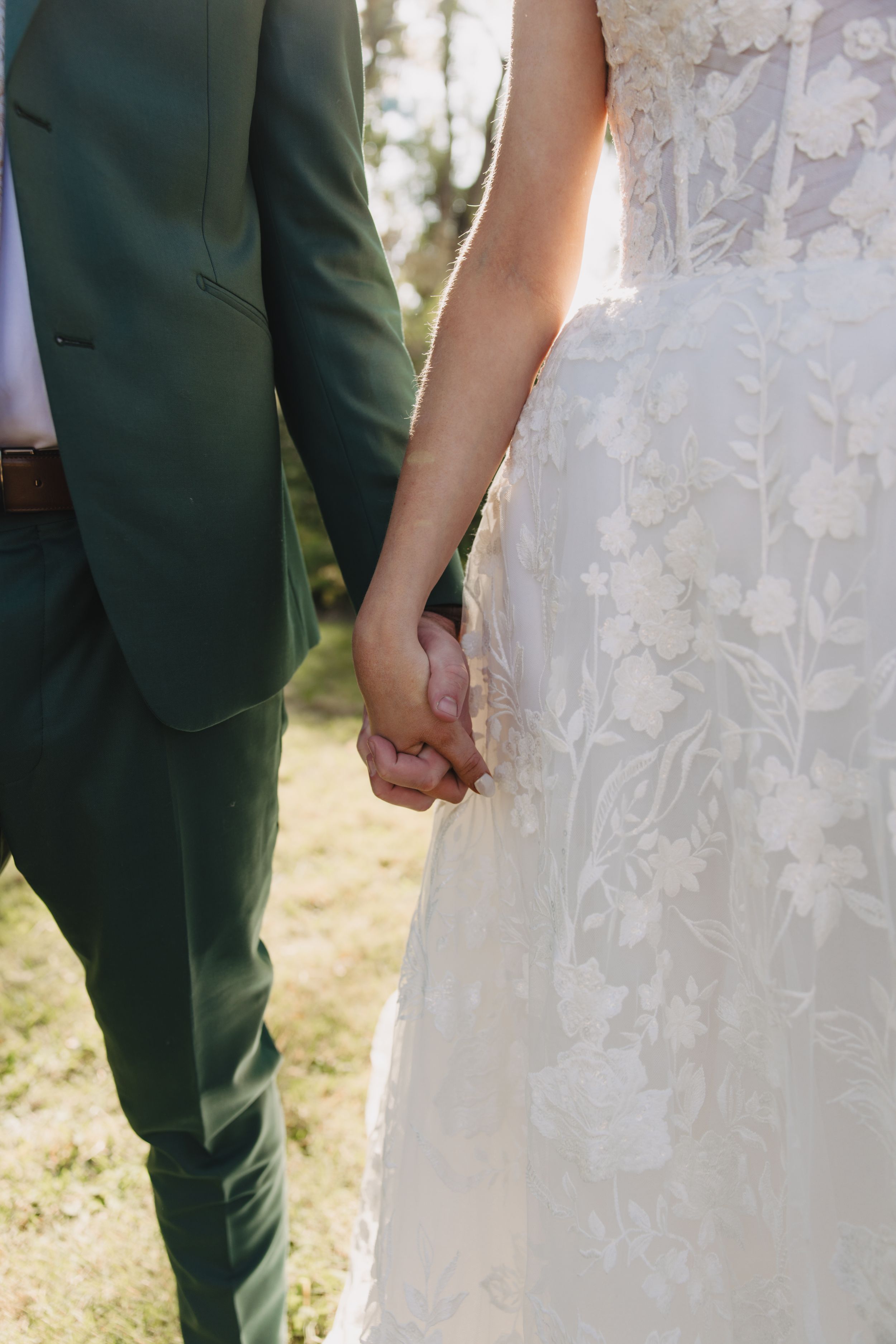A bride in a lace wedding dress stands next to her partner wearing a dark green suit as they hold hands outdoors.