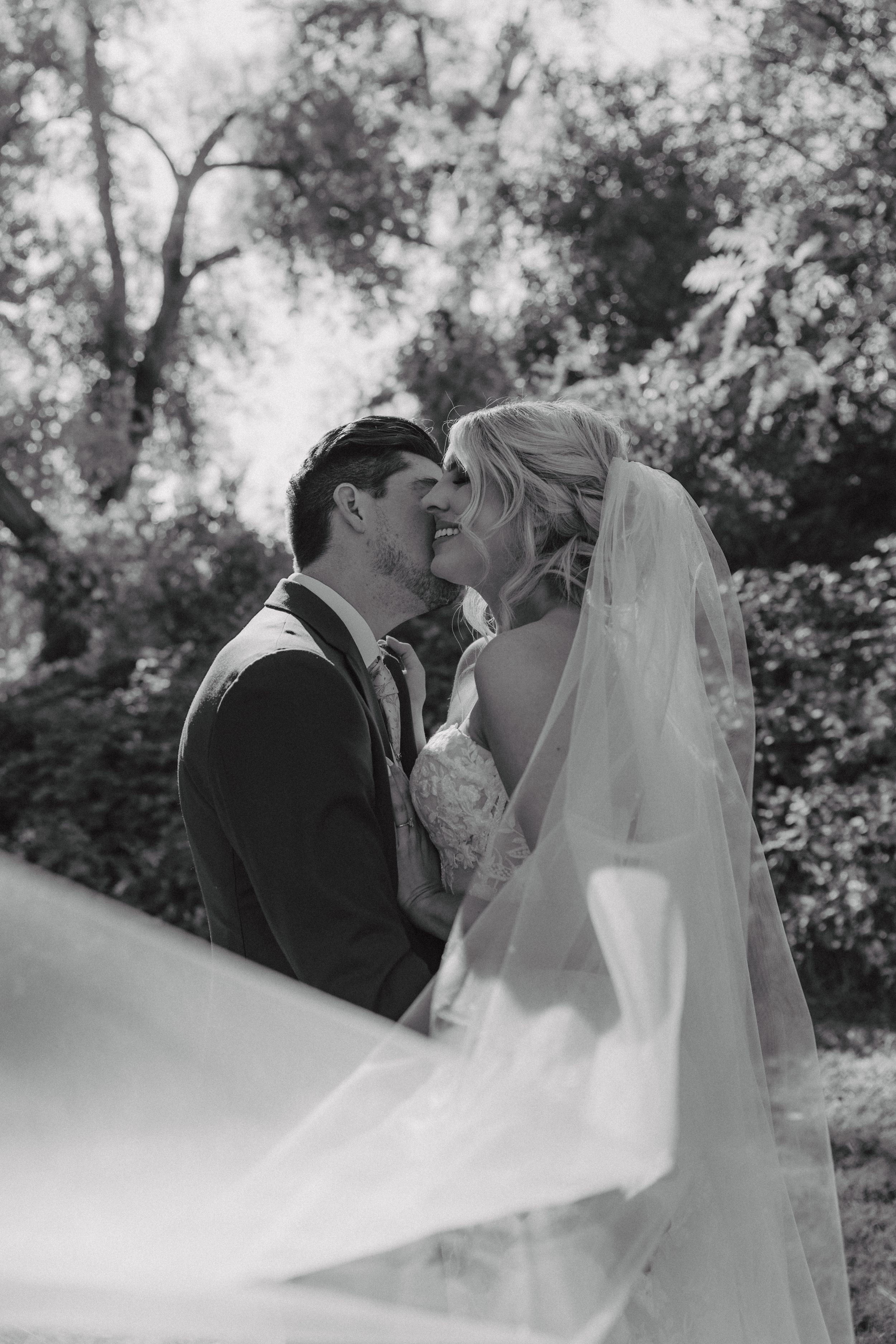 A wedding couple shares a romantic kiss outdoors surrounded by trees in black and white.
