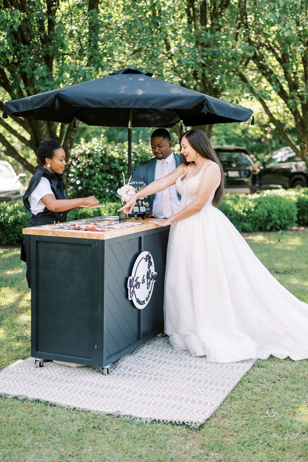 Elegant outdoor wedding bar setup with black counter and white decorative mat on grass in a garden setting.