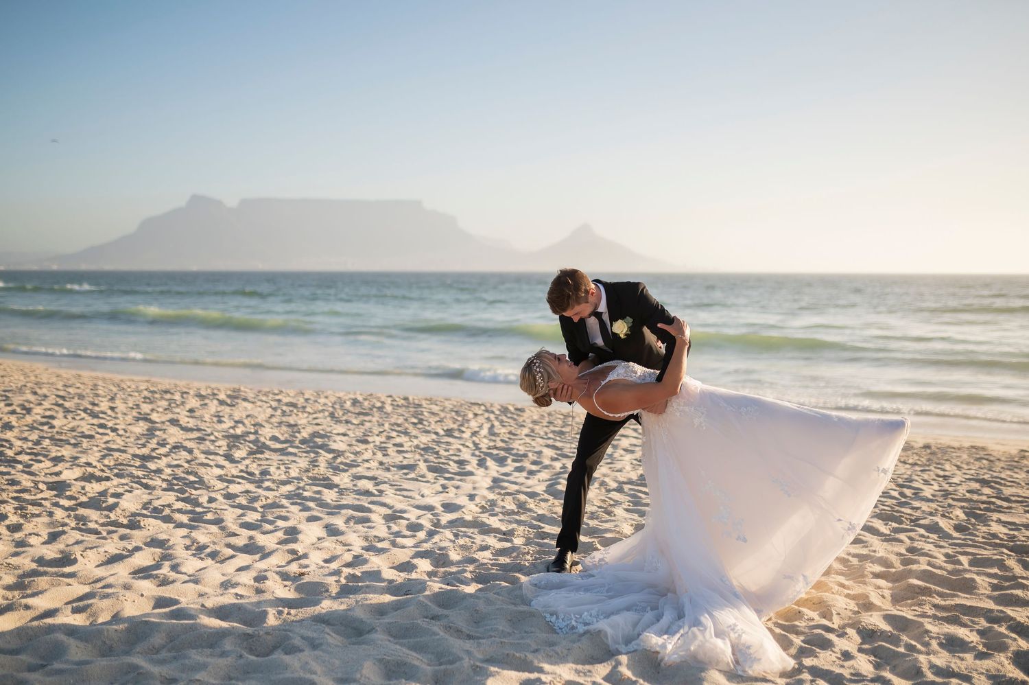 A series of professional wedding photos being taken on a sunny beach with mountain views and crashing waves in the background.