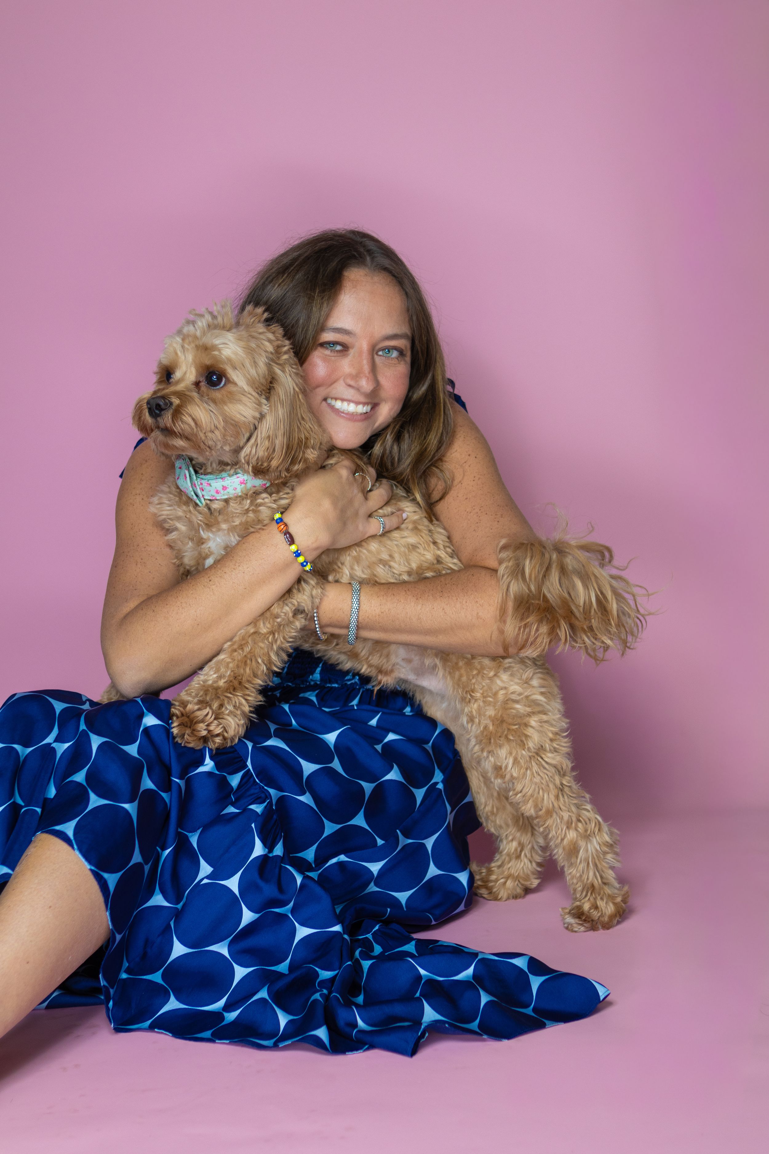 Smiling woman in blue polka dot dress hugging a fluffy golden doodle dog against a pink background.