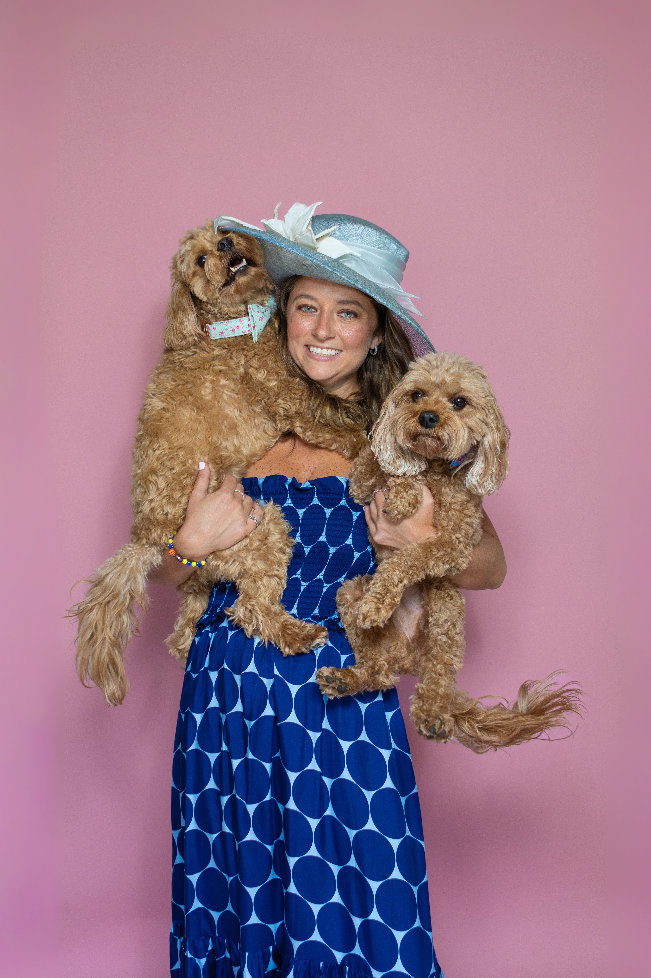 Woman in blue polka dot dress and hat holding two fluffy brown dogs against a pink background.