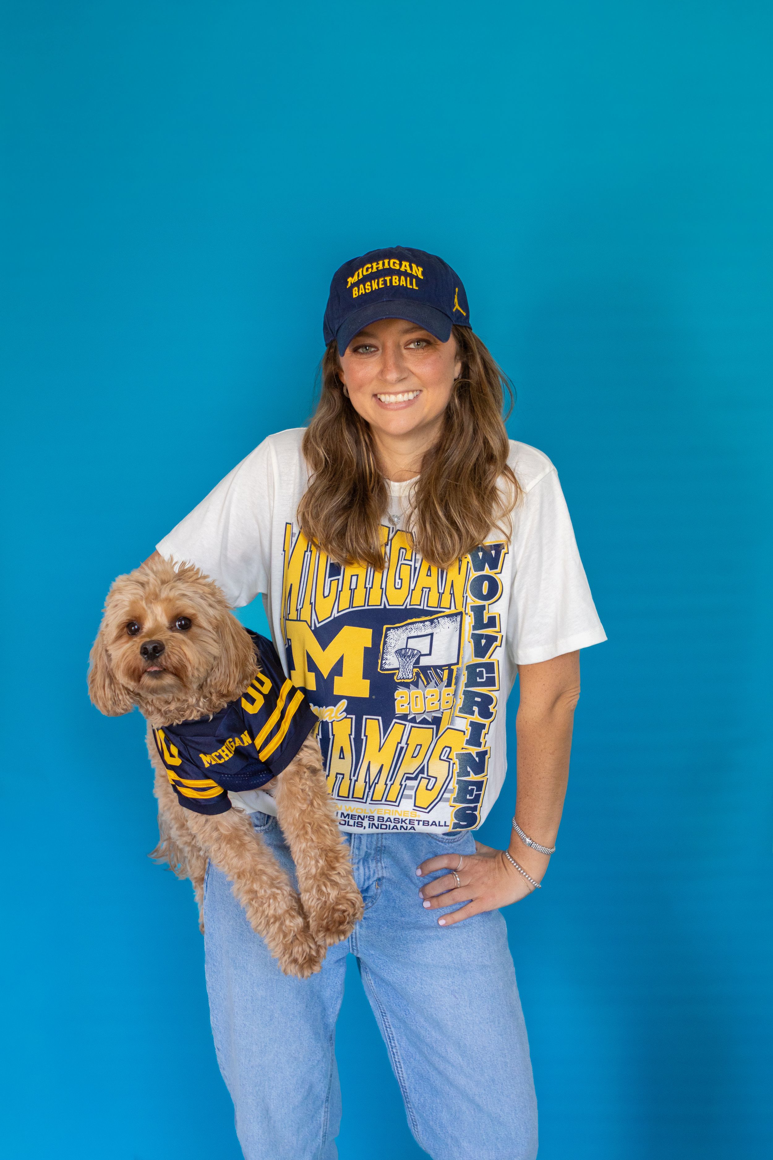 Woman in Michigan Wolverines basketball cap and tee holds a small dog wearing a matching Michigan jersey, blue background.