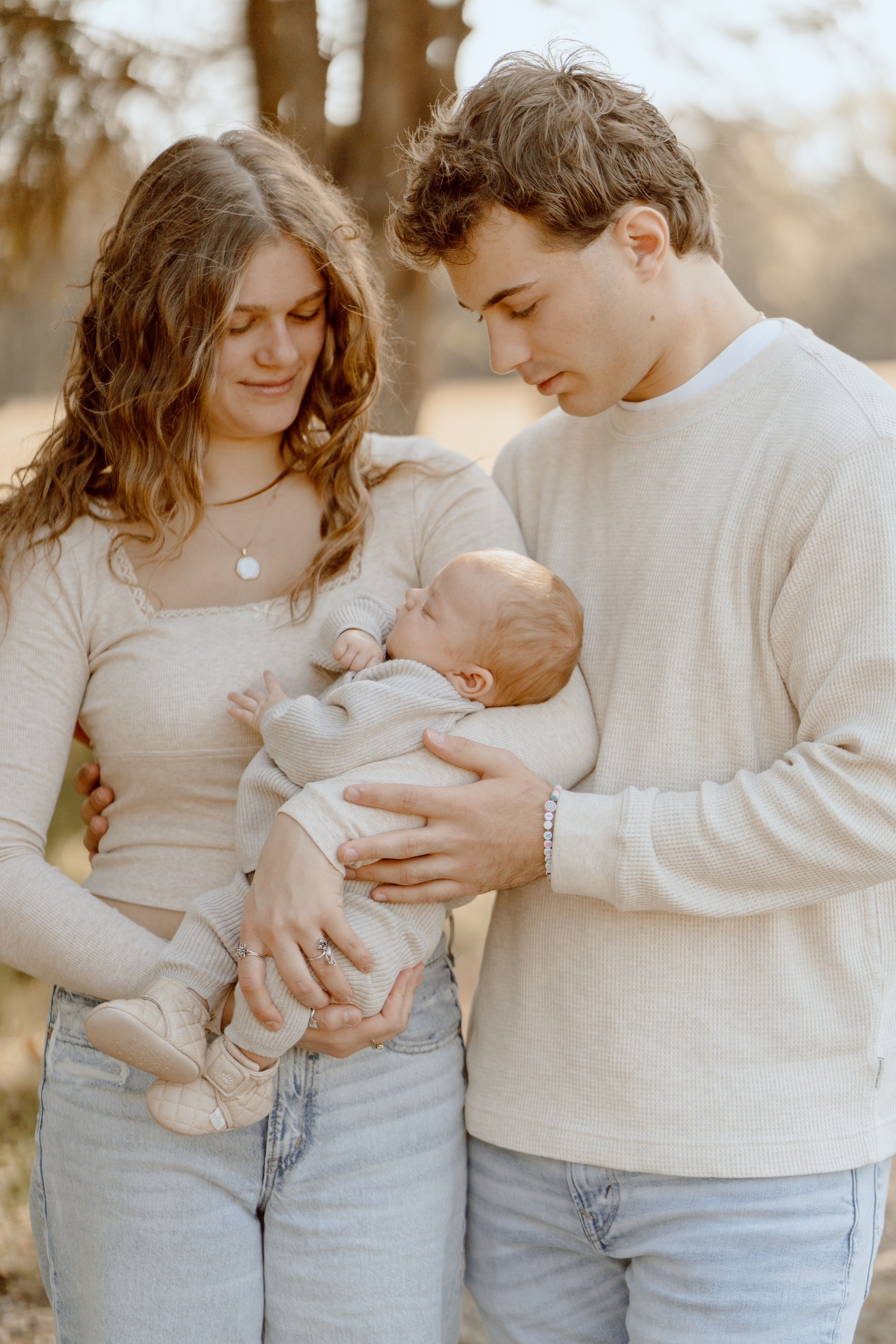 A family in cream-colored sweaters gazes lovingly at their newborn baby.