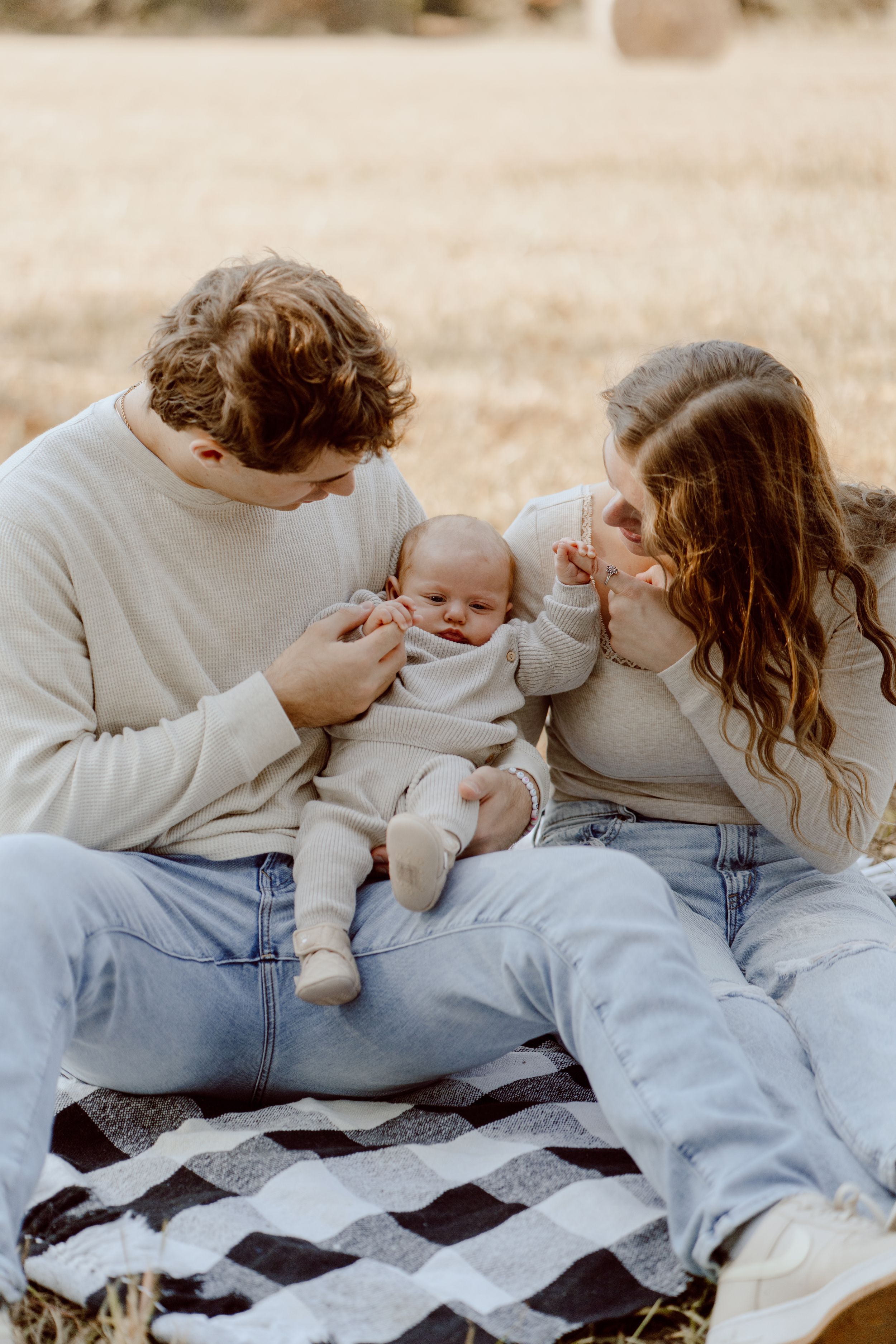 A family sits on a checkered blanket outdoors sharing a tender moment with their infant.