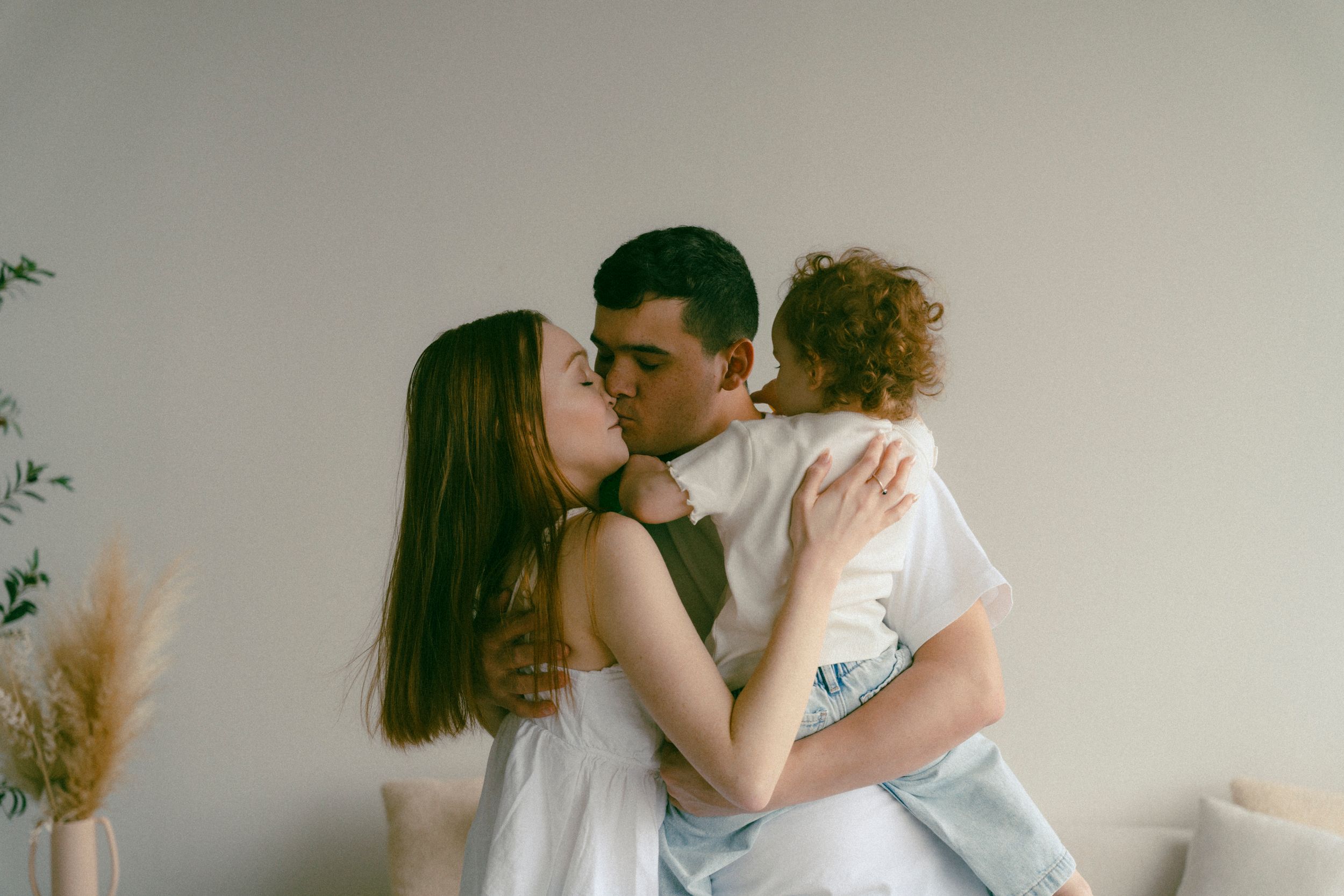 A young couple sharing a kiss while holding their curly-haired toddler in a bright, minimalist room.