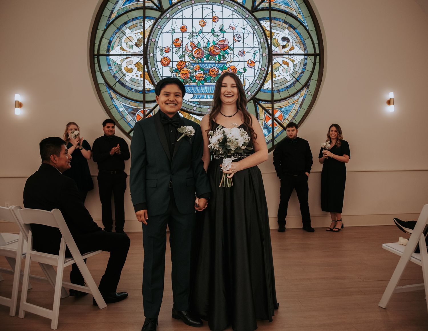 A formal couple stands in front of a stained glass window with wedding party members in black attire.