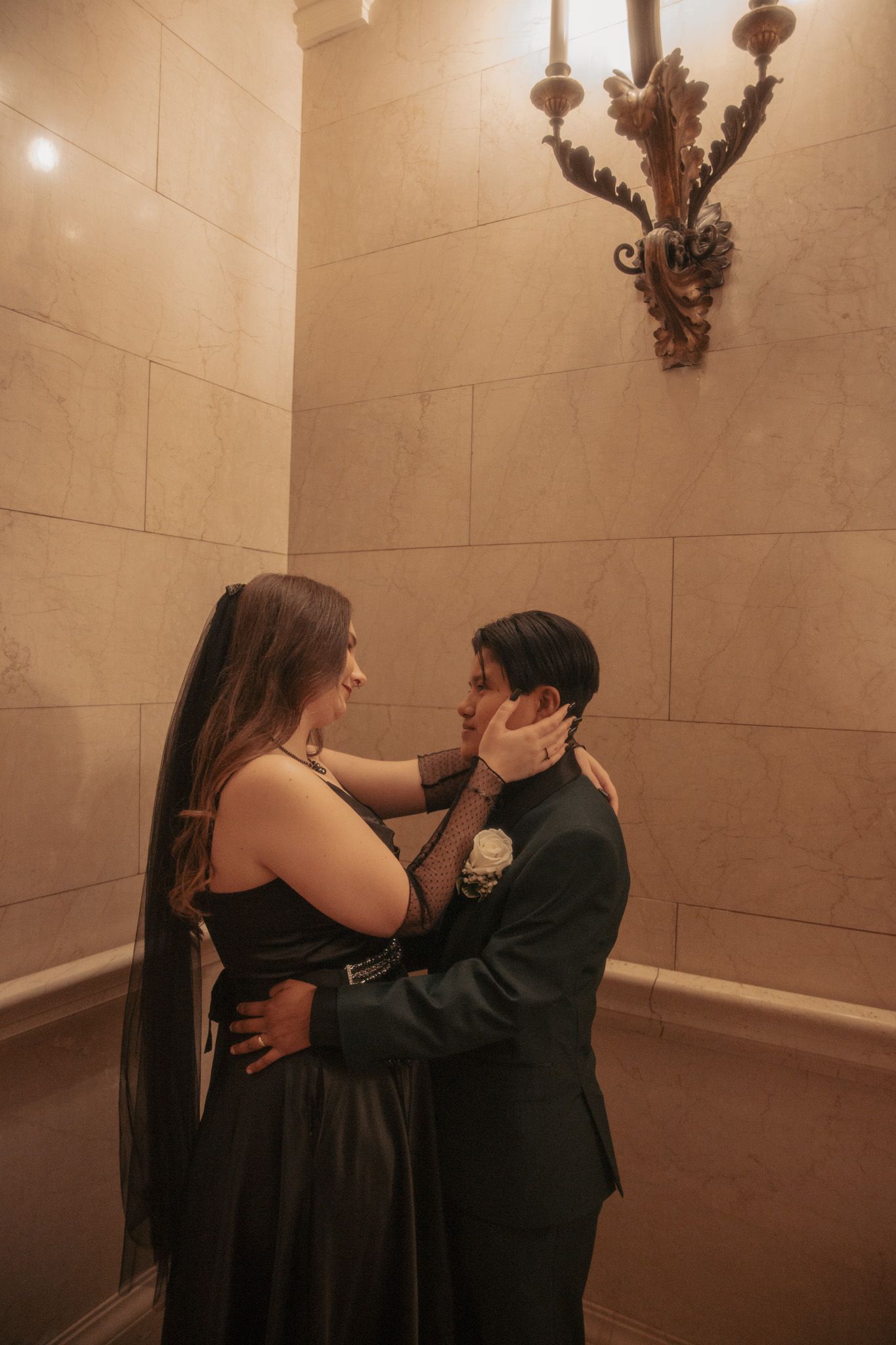 A couple in formal attire shares a tender moment in an elegant hallway with ornate wall sconce.