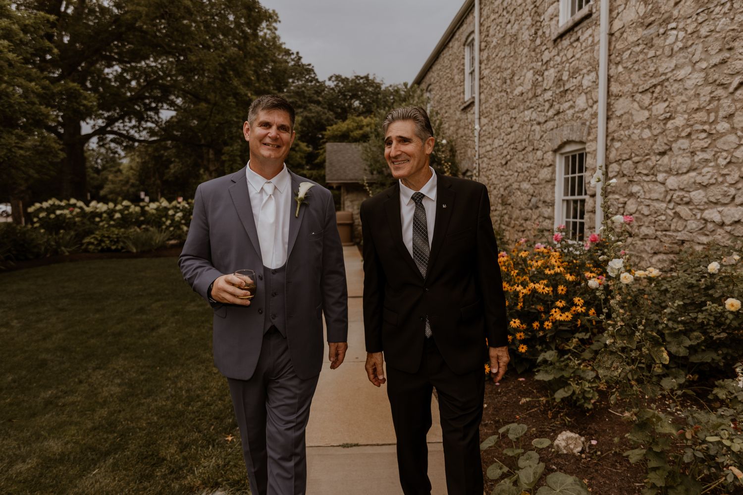 Wedding guests gather outside a rustic stone building on a sunny day.