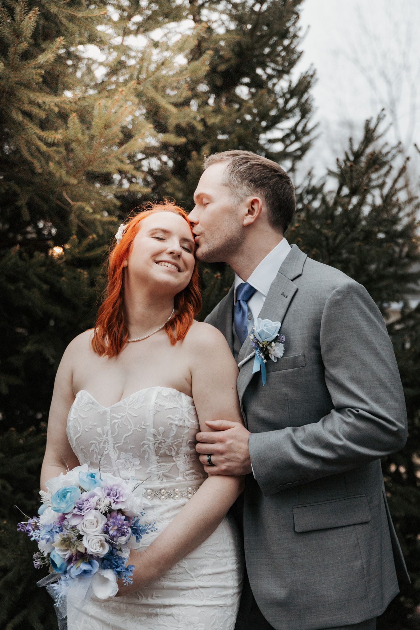 Newlyweds share tender moments outdoors with bride holding purple and blue floral bouquet.