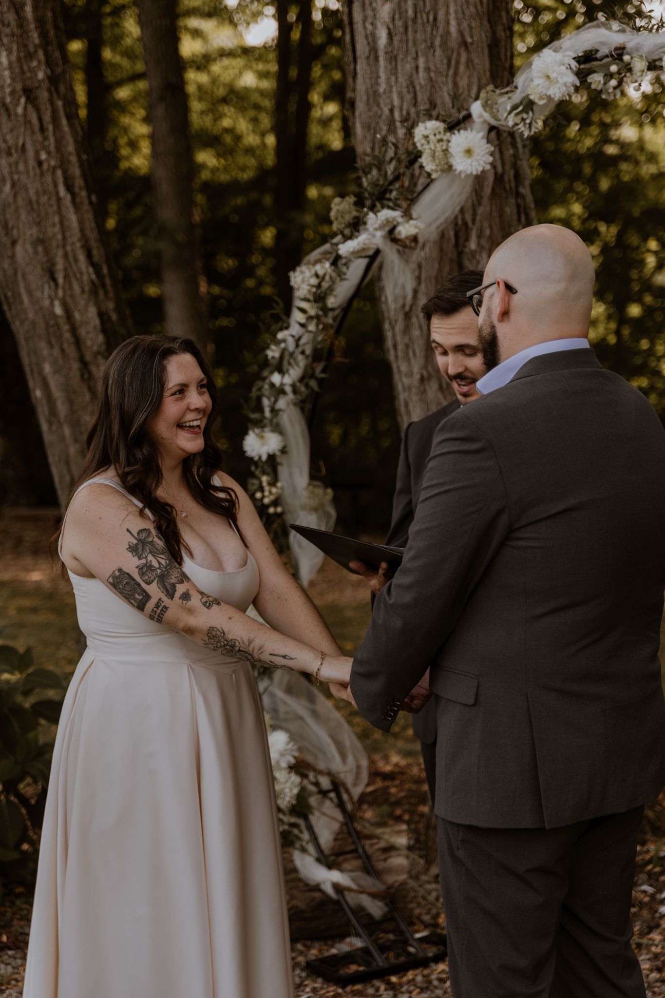 A romantic wedding ceremony takes place in a wooded setting with white floral decorations.
