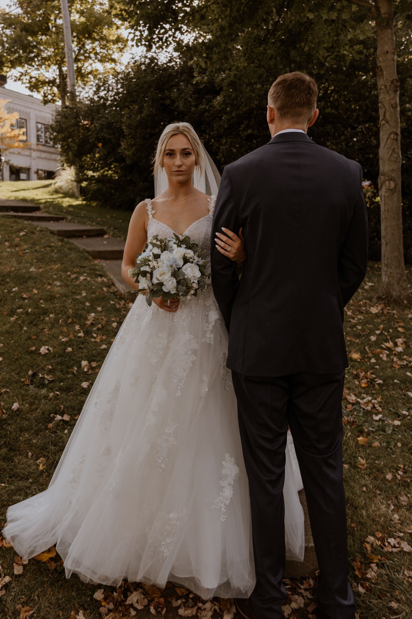 A bride in lace wedding gown holds bouquet while standing with groom in outdoor setting.