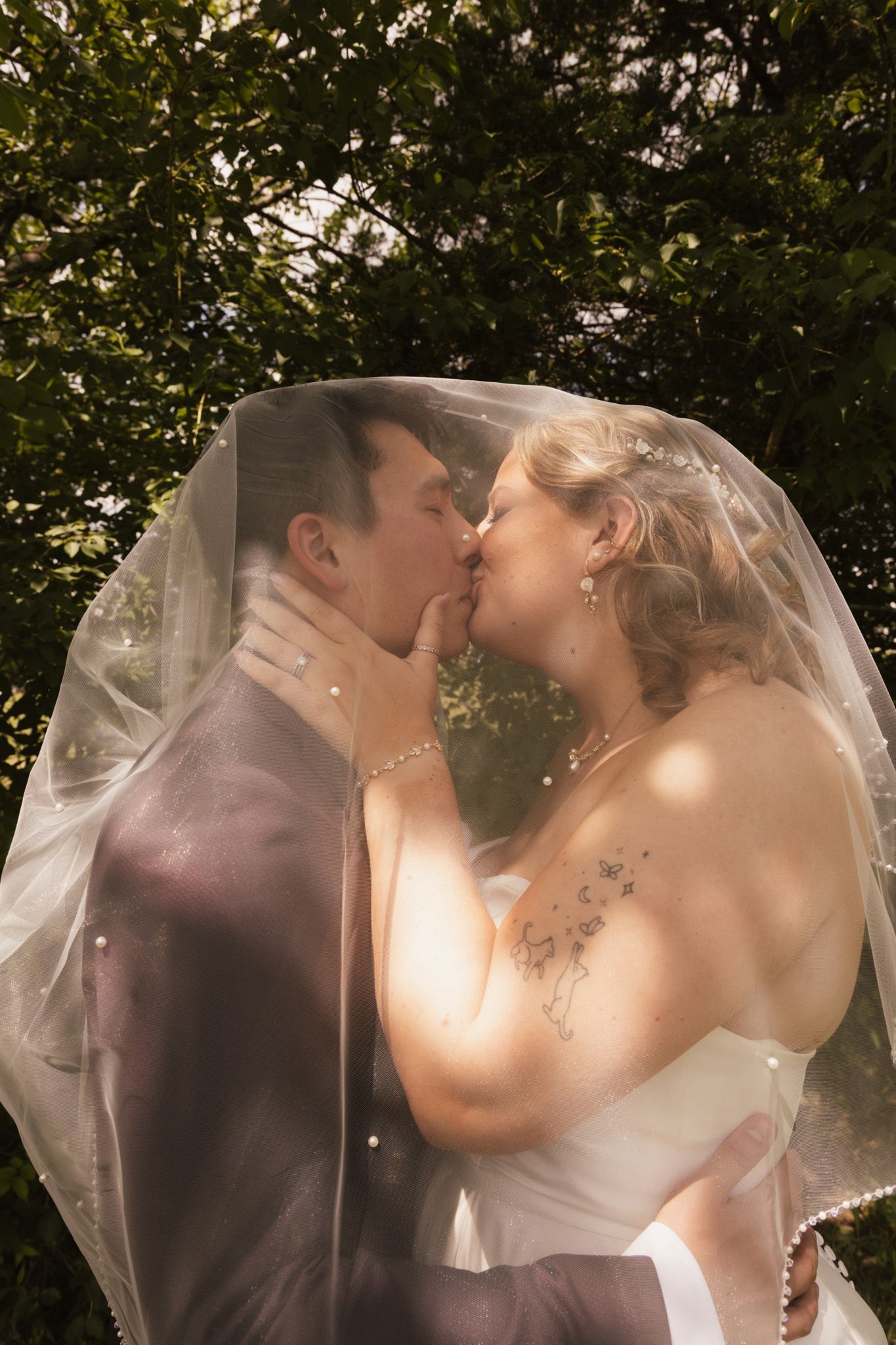 Newlyweds share tender kisses under a flowing veil in natural outdoor lighting.