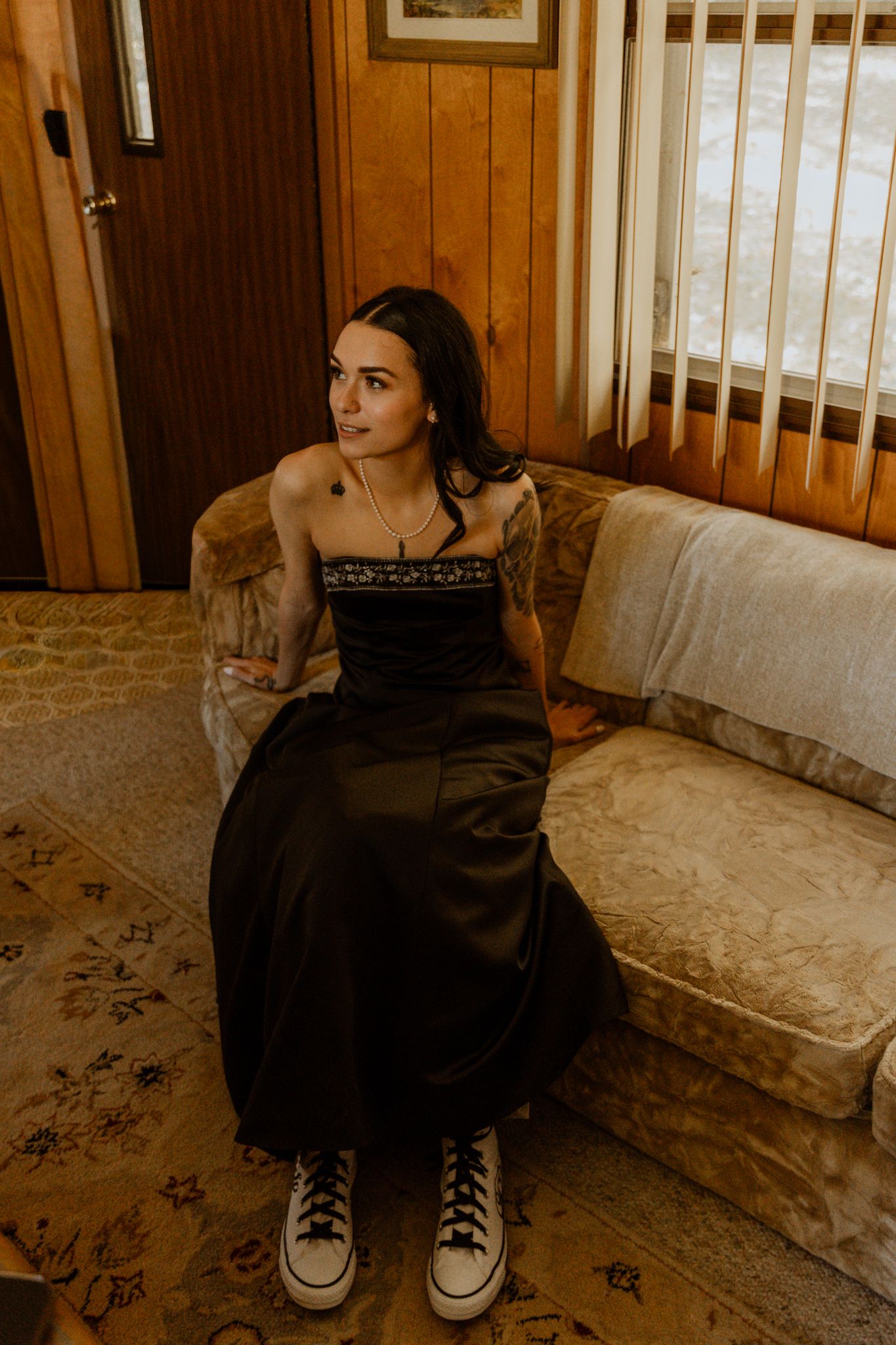 A person in black attire poses on a vintage couch in a wood-paneled room.