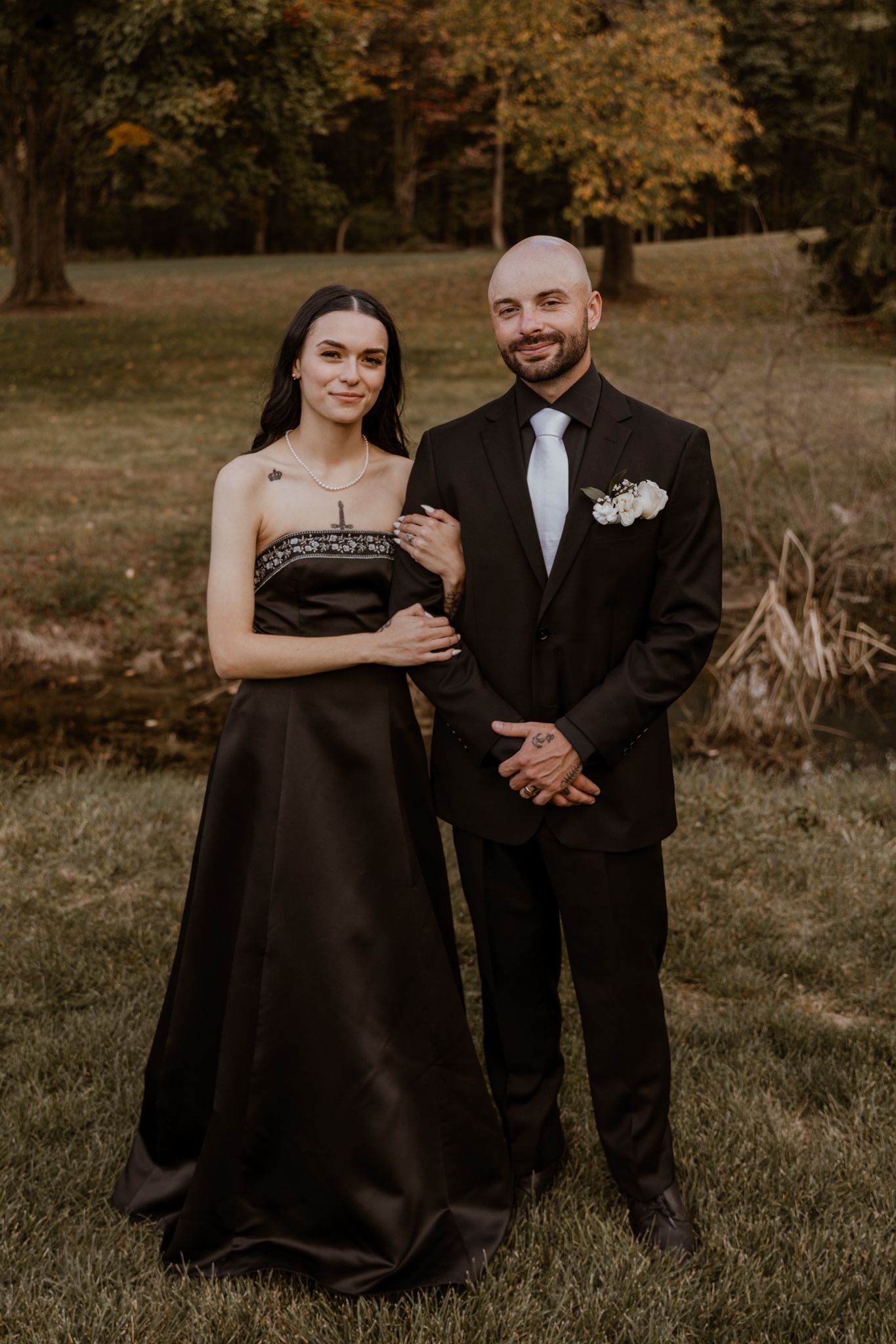 A couple in formal black attire poses together on grassy grounds during autumn.
