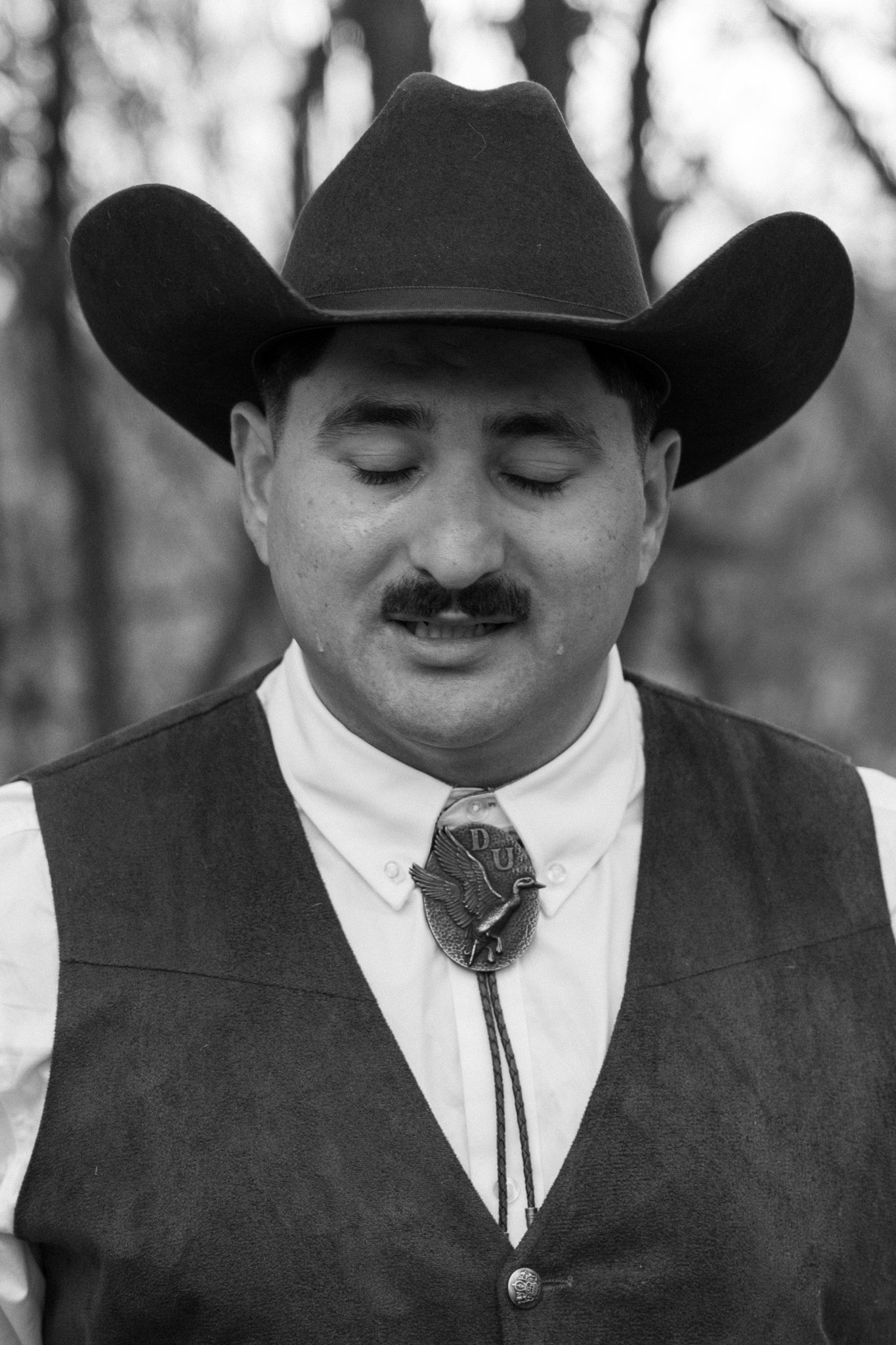 Black and white portrait of a man in a cowboy hat, vest, and bolo tie with eyes closed outdoors.