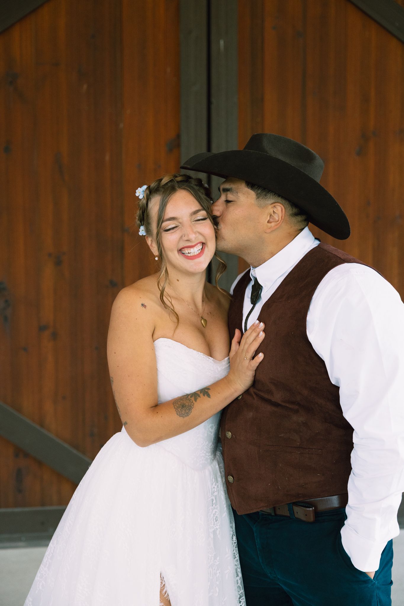 A groom in a cowboy hat kisses his laughing bride in a white strapless gown near rustic wooden barn doors.