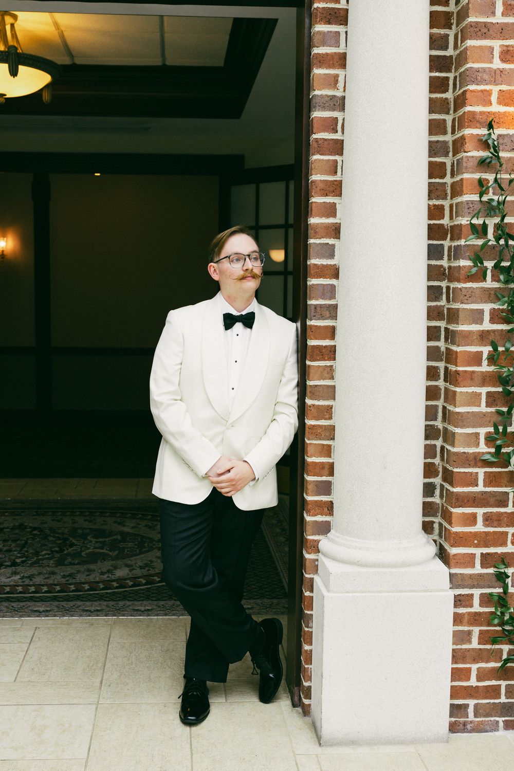 Groom in white tuxedo jacket and black bow tie leaning against brick column at wedding venue.