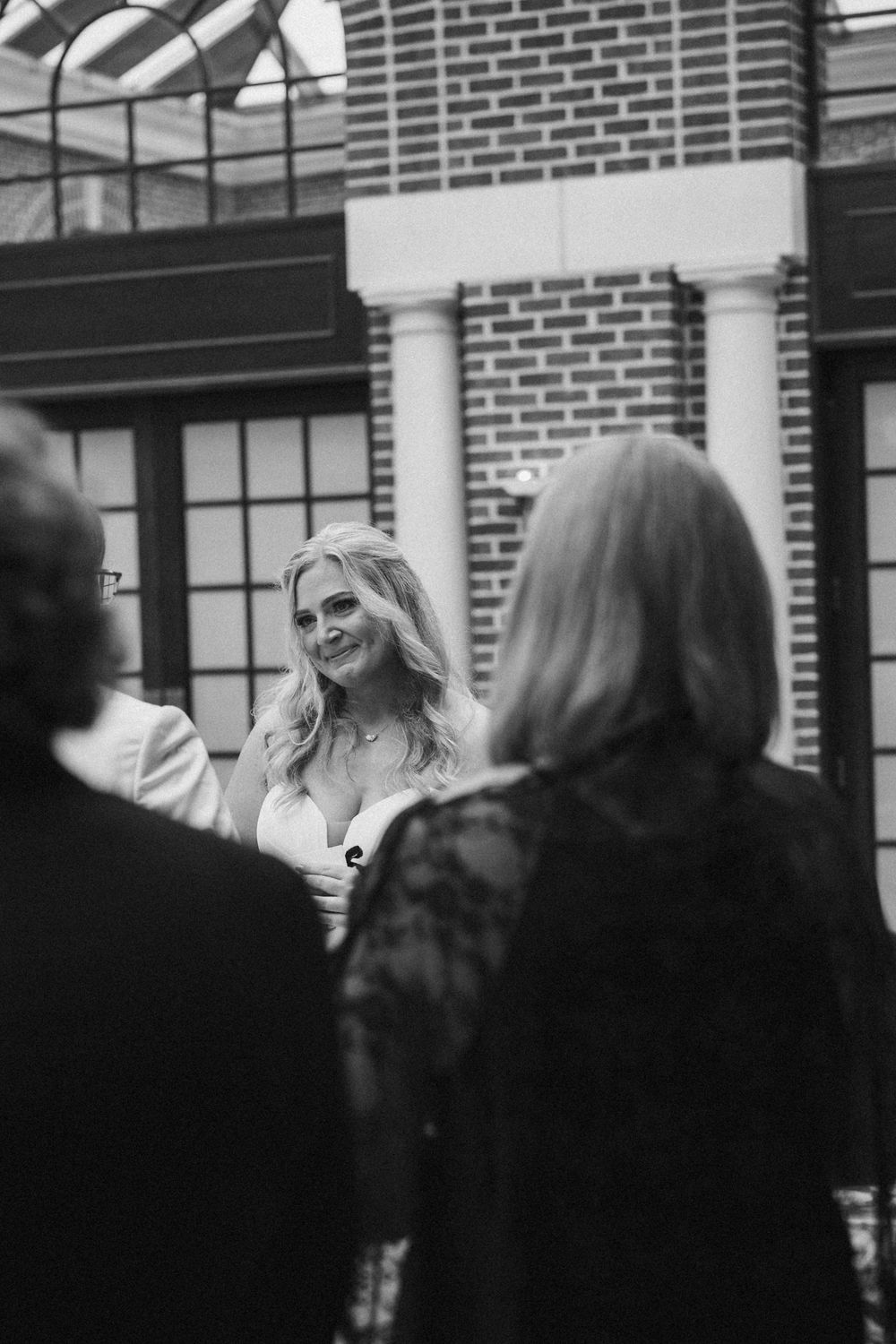 Black and white photo of a smiling blonde bride in a wedding dress greeting guests outside a brick building.