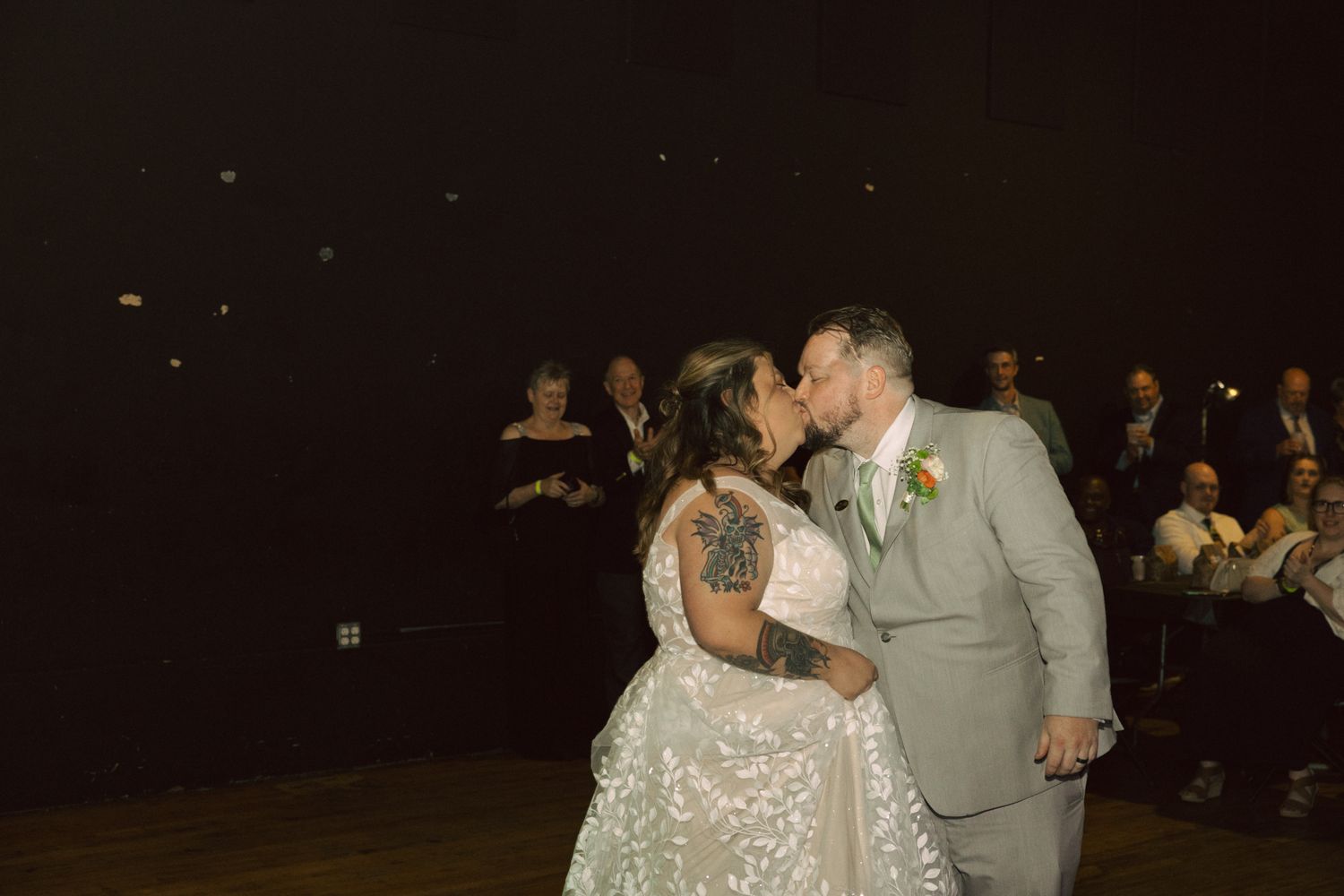 Bride and groom share a first kiss on the dance floor at their wedding reception, guests seated in background.