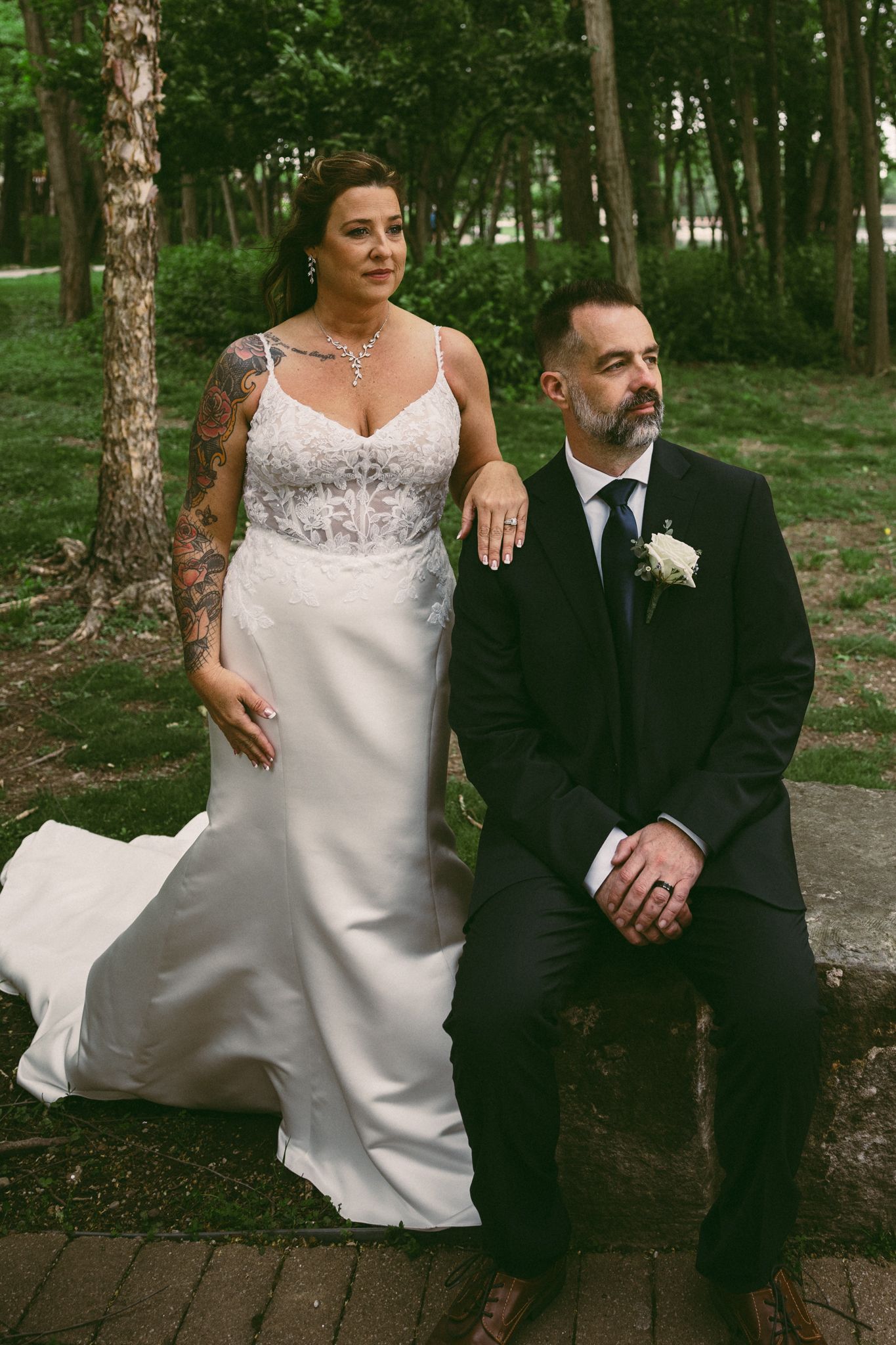 Bride in white lace gown with tattoo stands beside groom in black suit with boutonniere in a park setting.