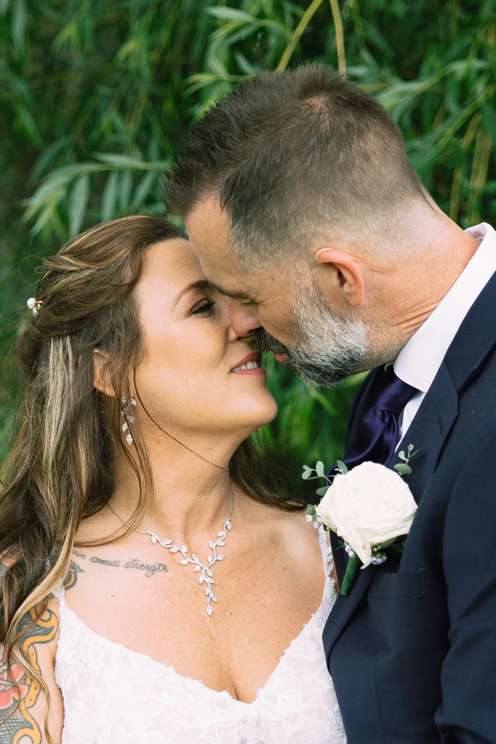 A bride and groom share an intimate moment, touching foreheads, with greenery in the background at their wedding.