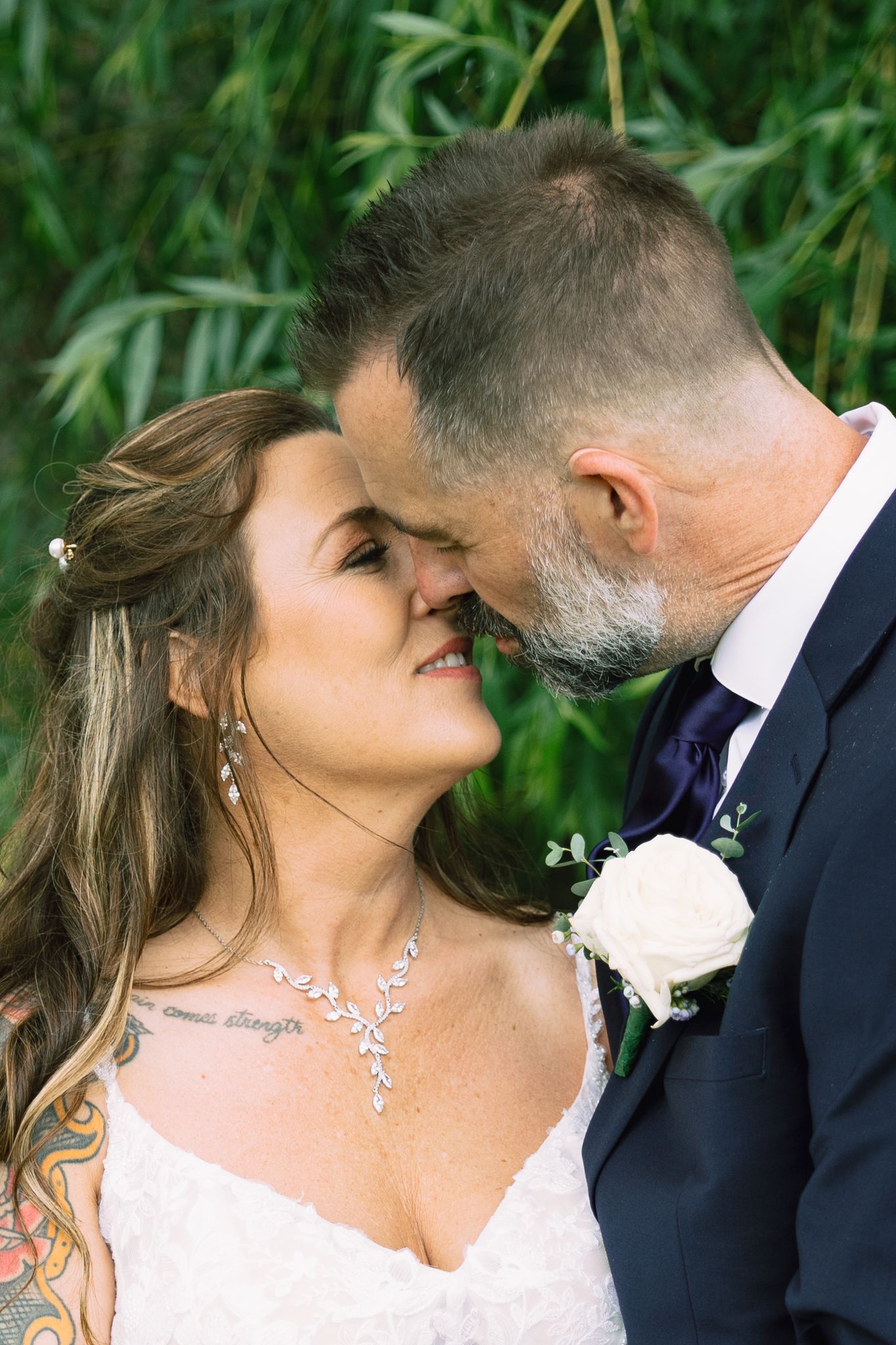 A bride and groom share an intimate moment, touching foreheads, with greenery in the background at their wedding.