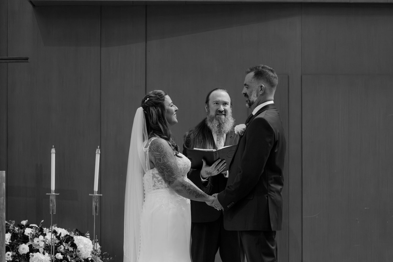 Black and white photo of a bride and groom holding hands during wedding ceremony with officiant behind them.