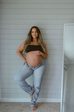 Casual maternity photo against a white wall showing a pregnant woman in comfortable clothing.