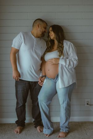 Barefoot couple poses against white brick wall in casual maternity photos.