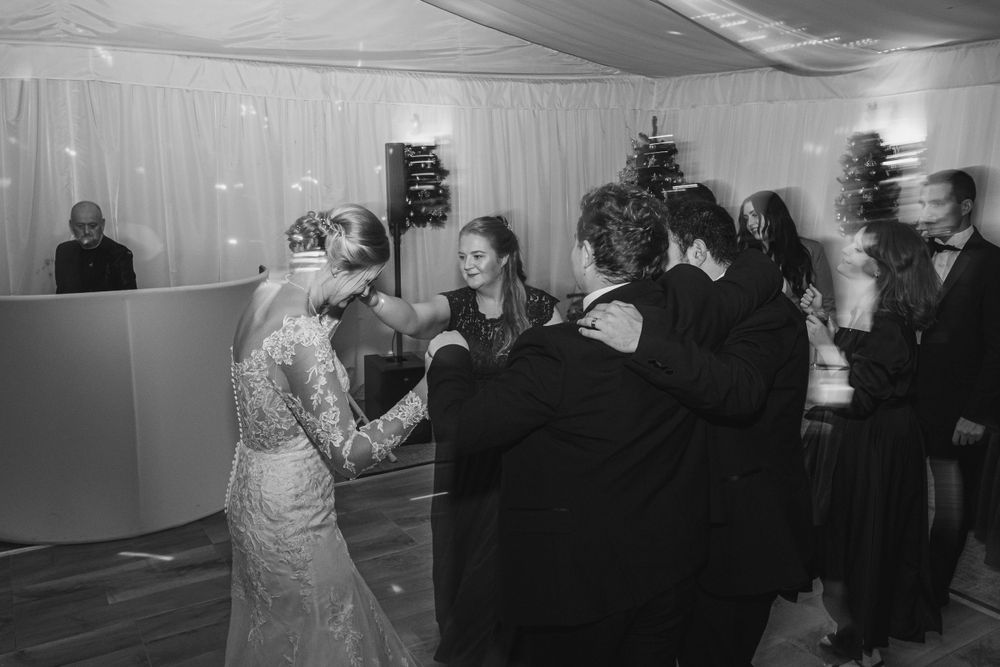 Wedding guests dance in a line formation at an indoor reception venue with white drapes.