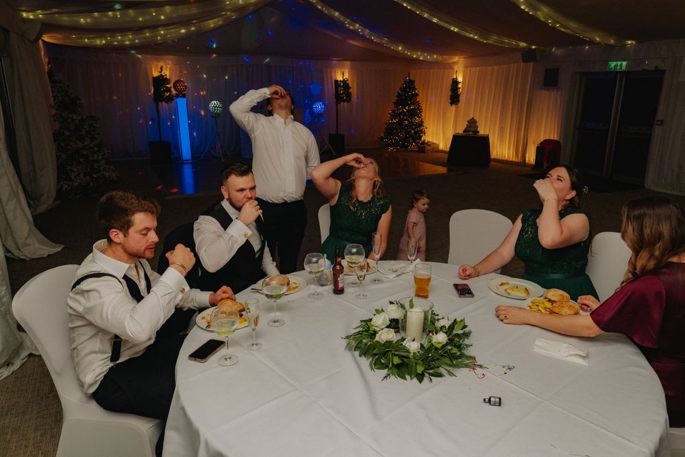 Wedding reception guests enjoying dinner at decorated round tables with centerpieces and mood lighting.