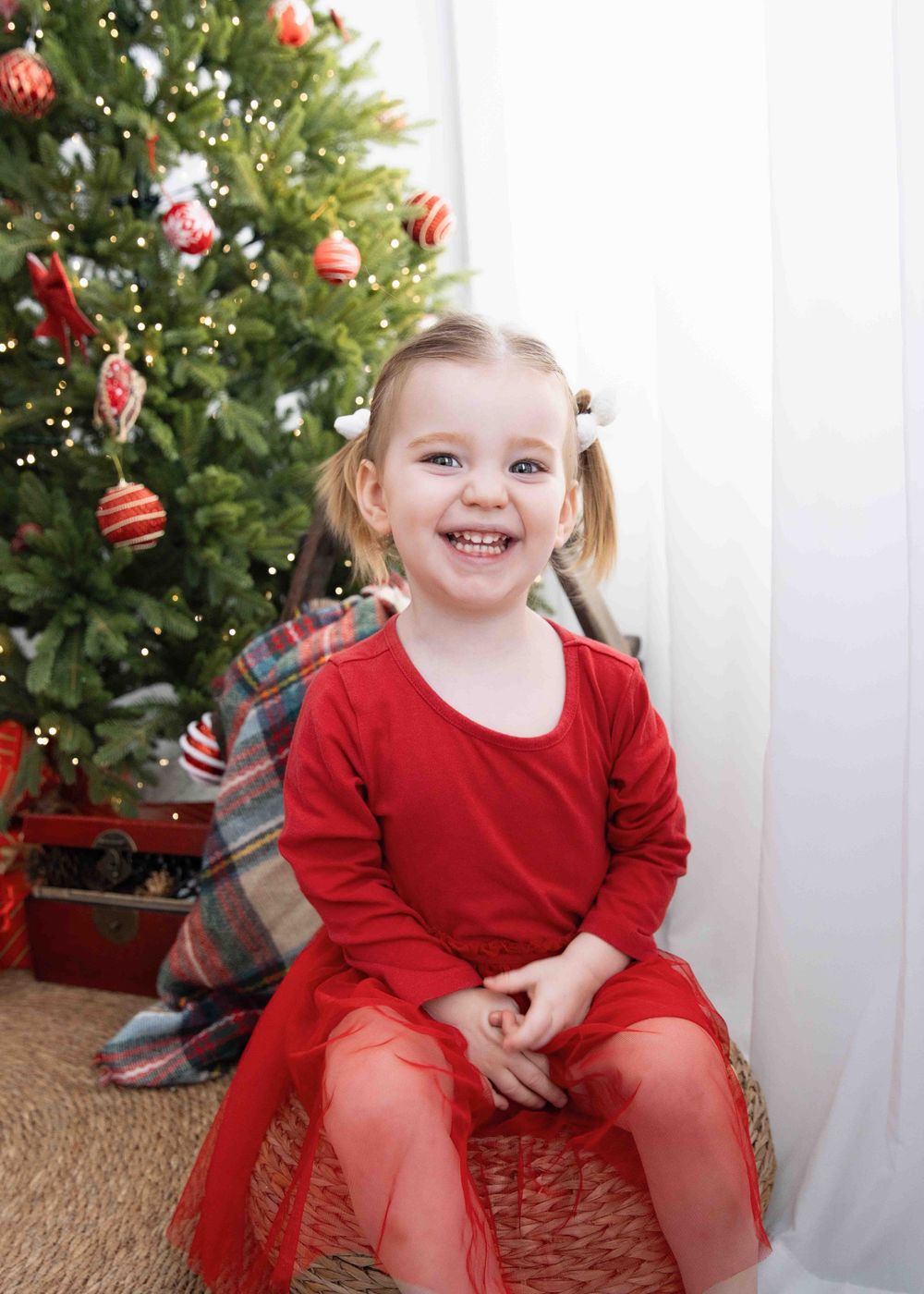 A child in a red top and tulle skirt sits near a decorated Christmas tree with ornaments and lights. Lullaby Images Christmas Mini Photos