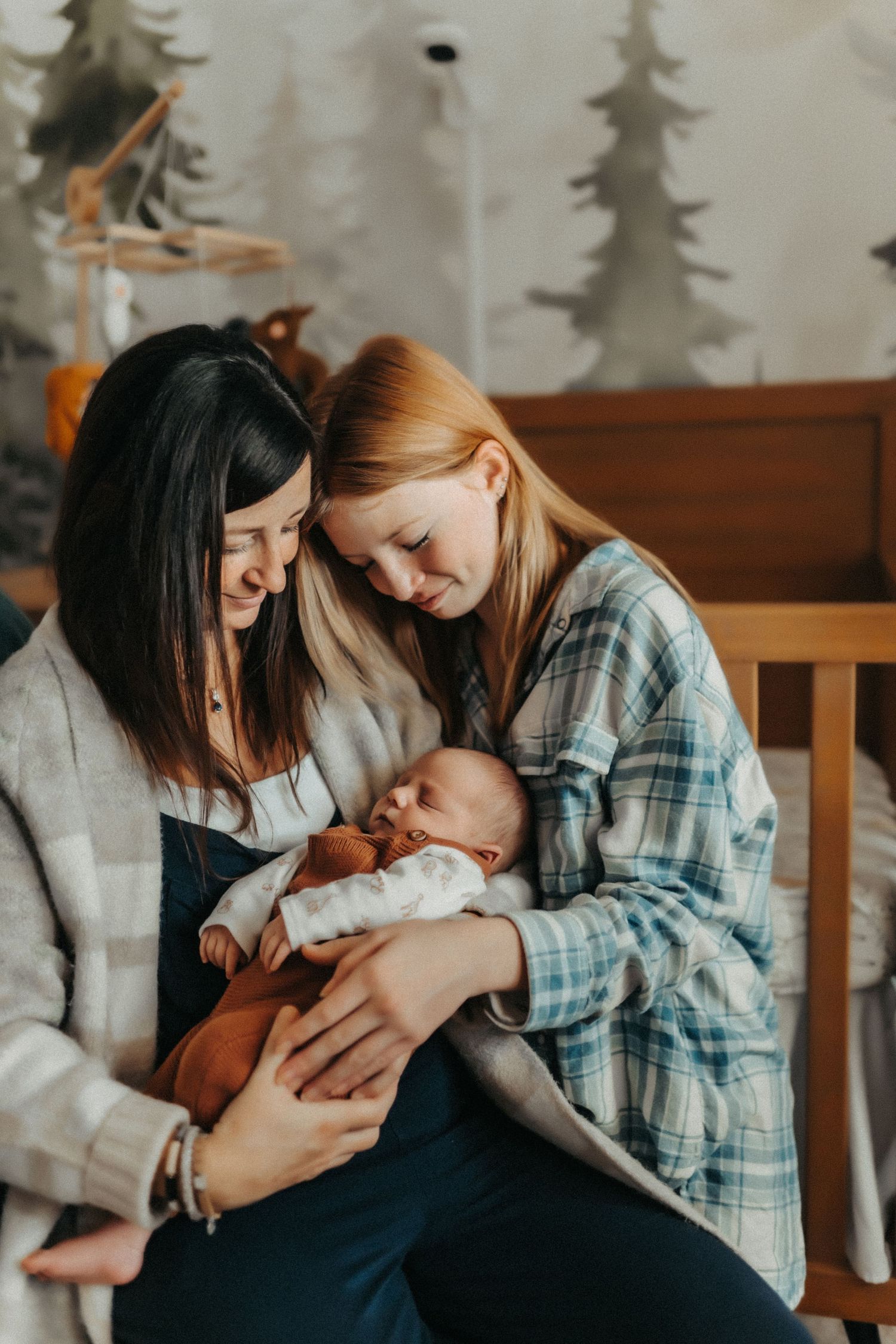 Two people sharing a tender moment with a sleeping baby in their arms on a wooden bench with pine trees in background.