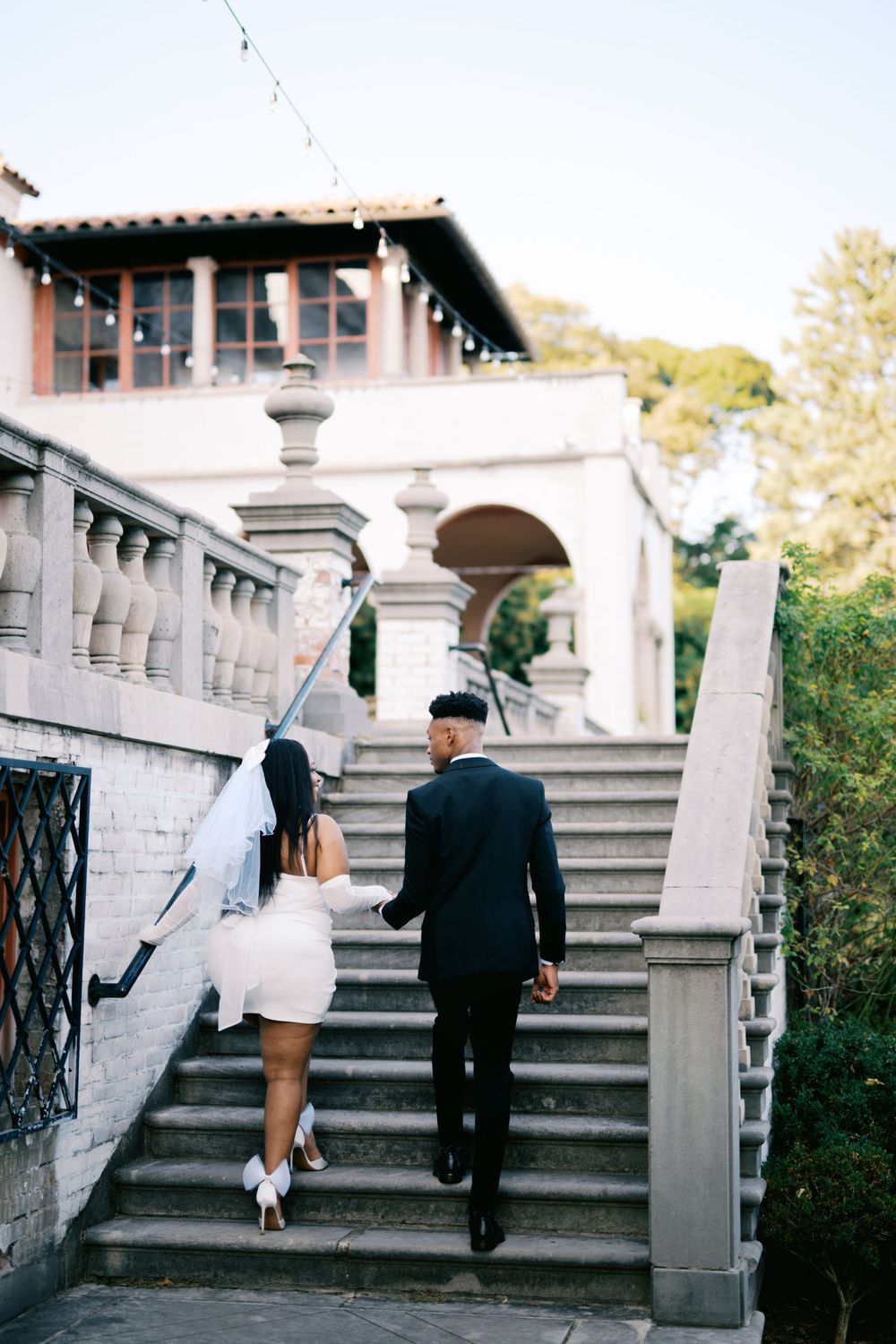 A couple in wedding attire walking up an elegant stone staircase at an outdoor venue with Mediterranean architecture.
