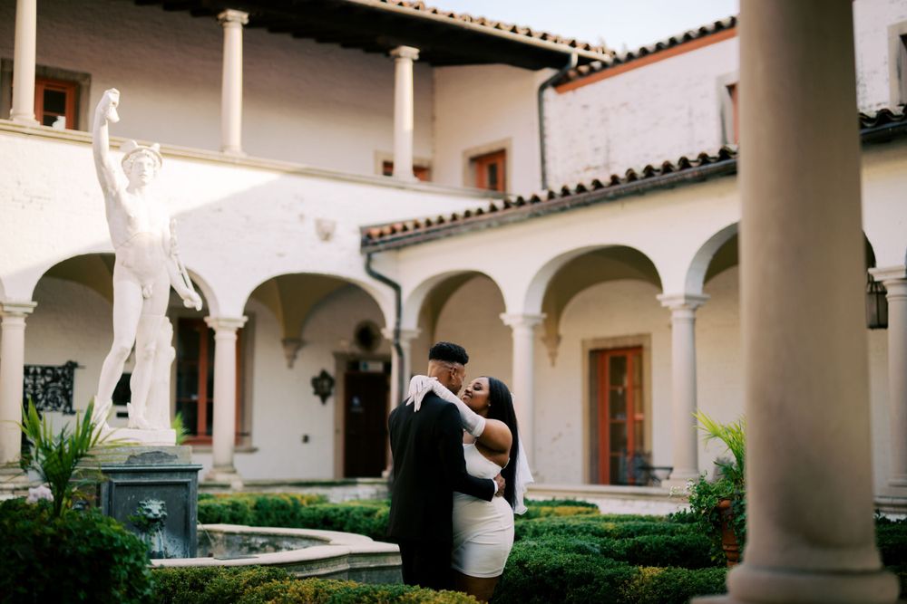 A couple shares an intimate moment in a Spanish colonial courtyard with white arches and tiled roof architecture.